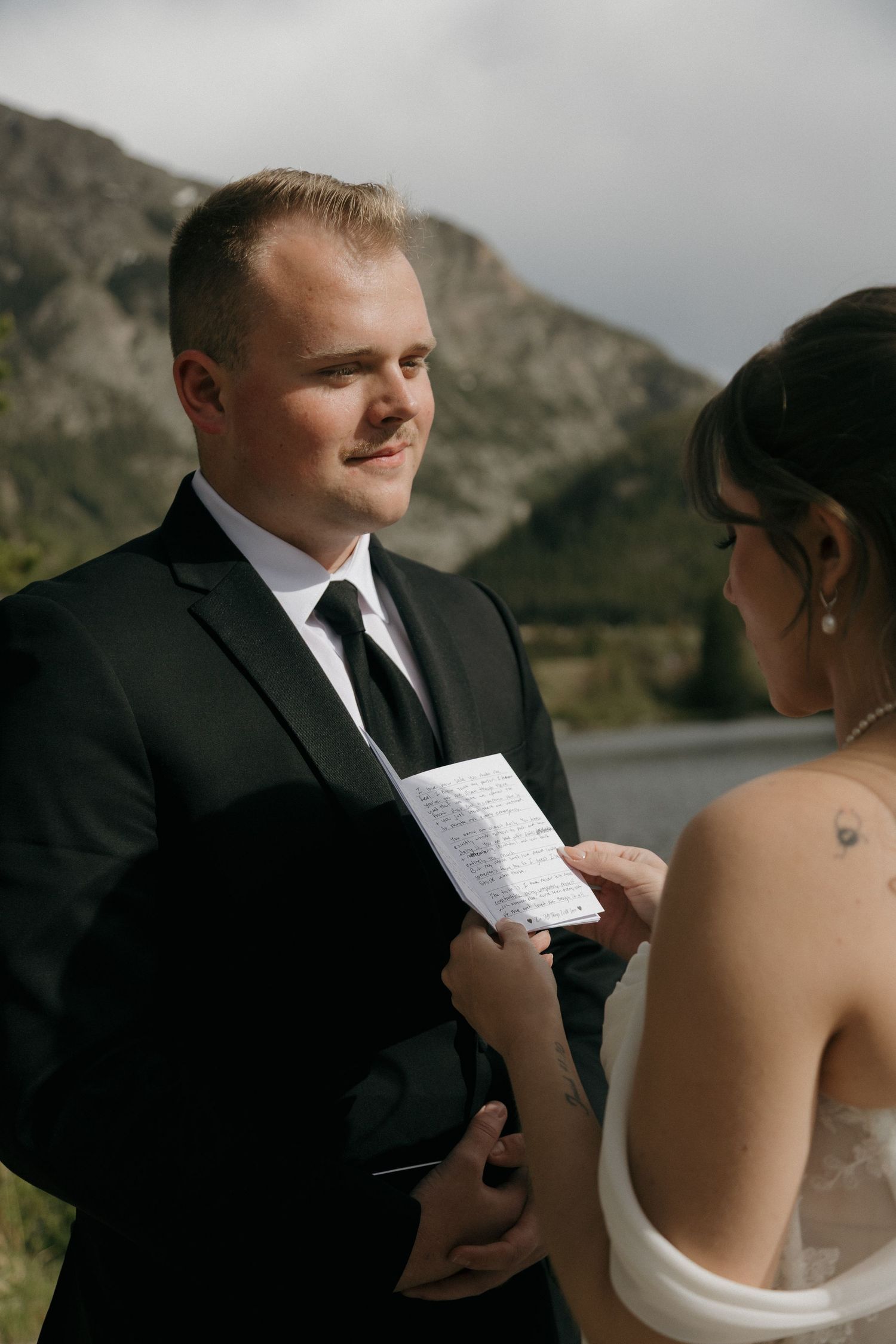 Groom in black suit reads wedding vows to bride outdoors with mountain lake backdrop.