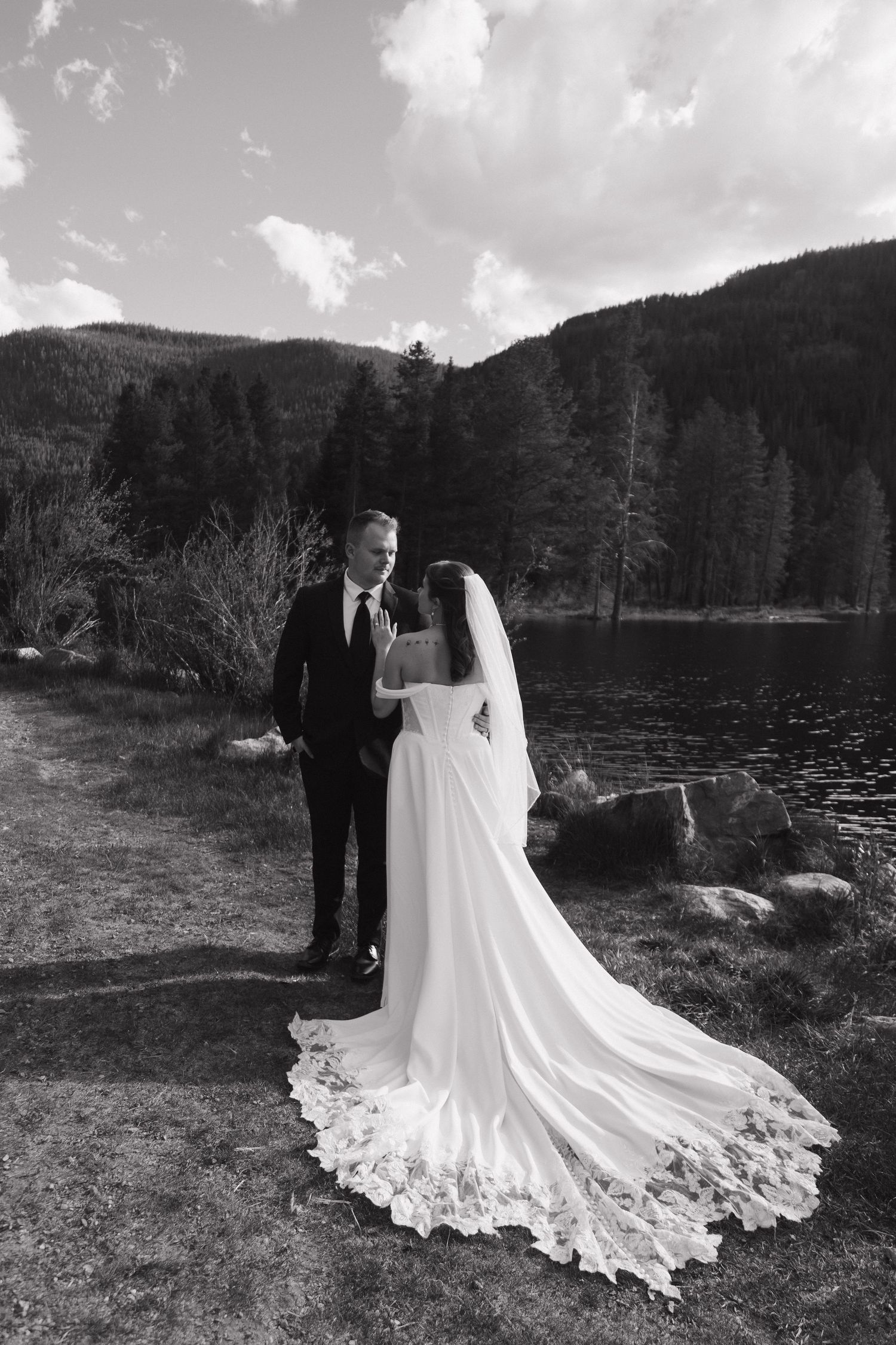 Black and white portrait of bride and groom standing together near a mountain lake with lace veil train.