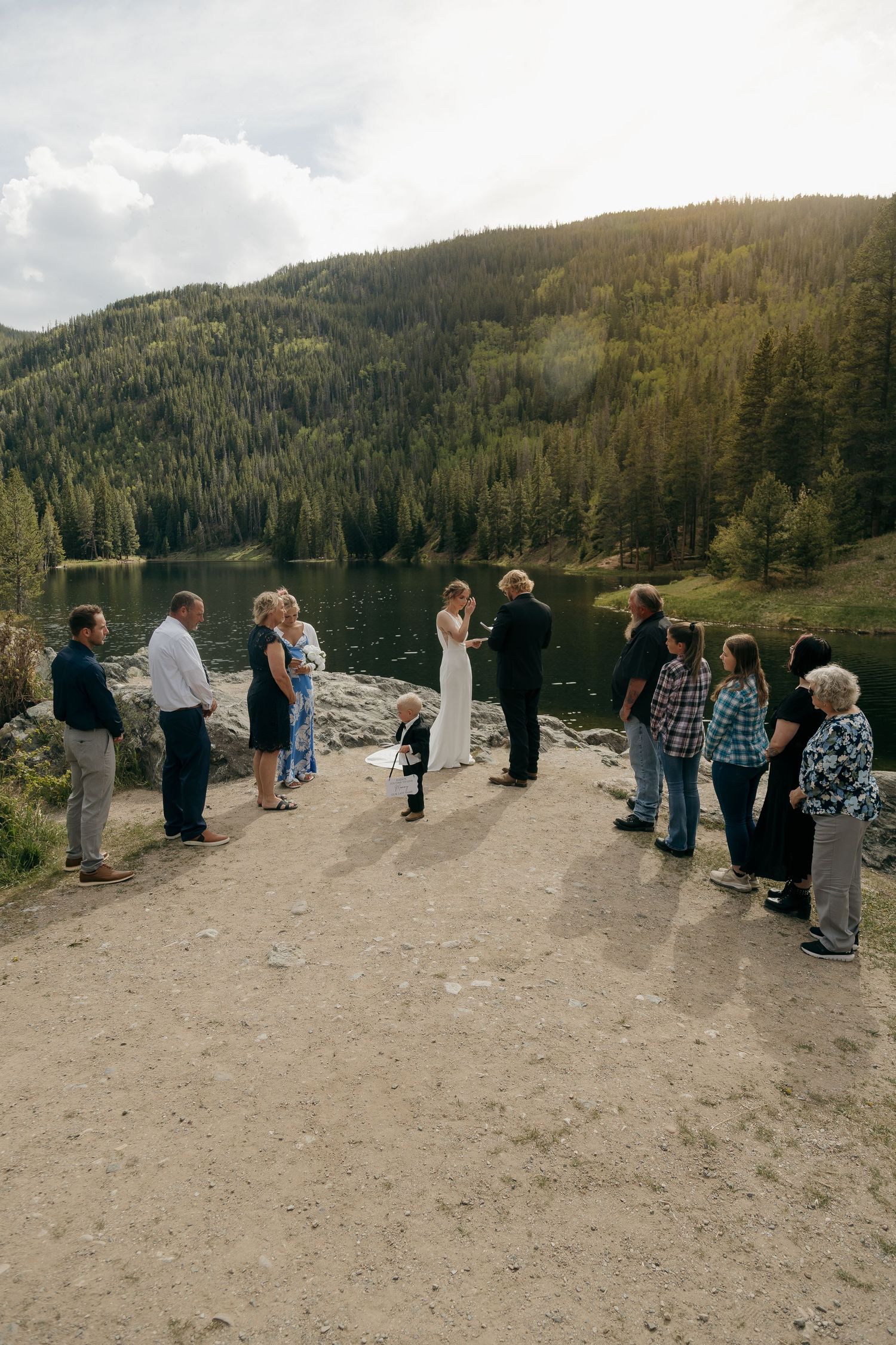 Small outdoor wedding ceremony by a mountain lake surrounded by evergreen forests with guests standing in a circle.