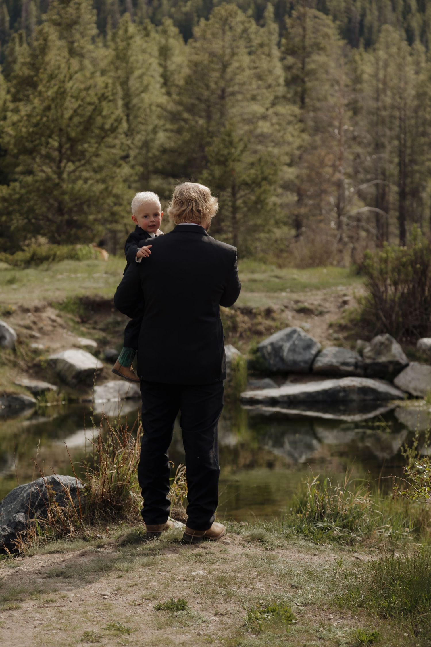 A man in a dark suit holds a young blonde child while standing near a mountain lake surrounded by pine trees.