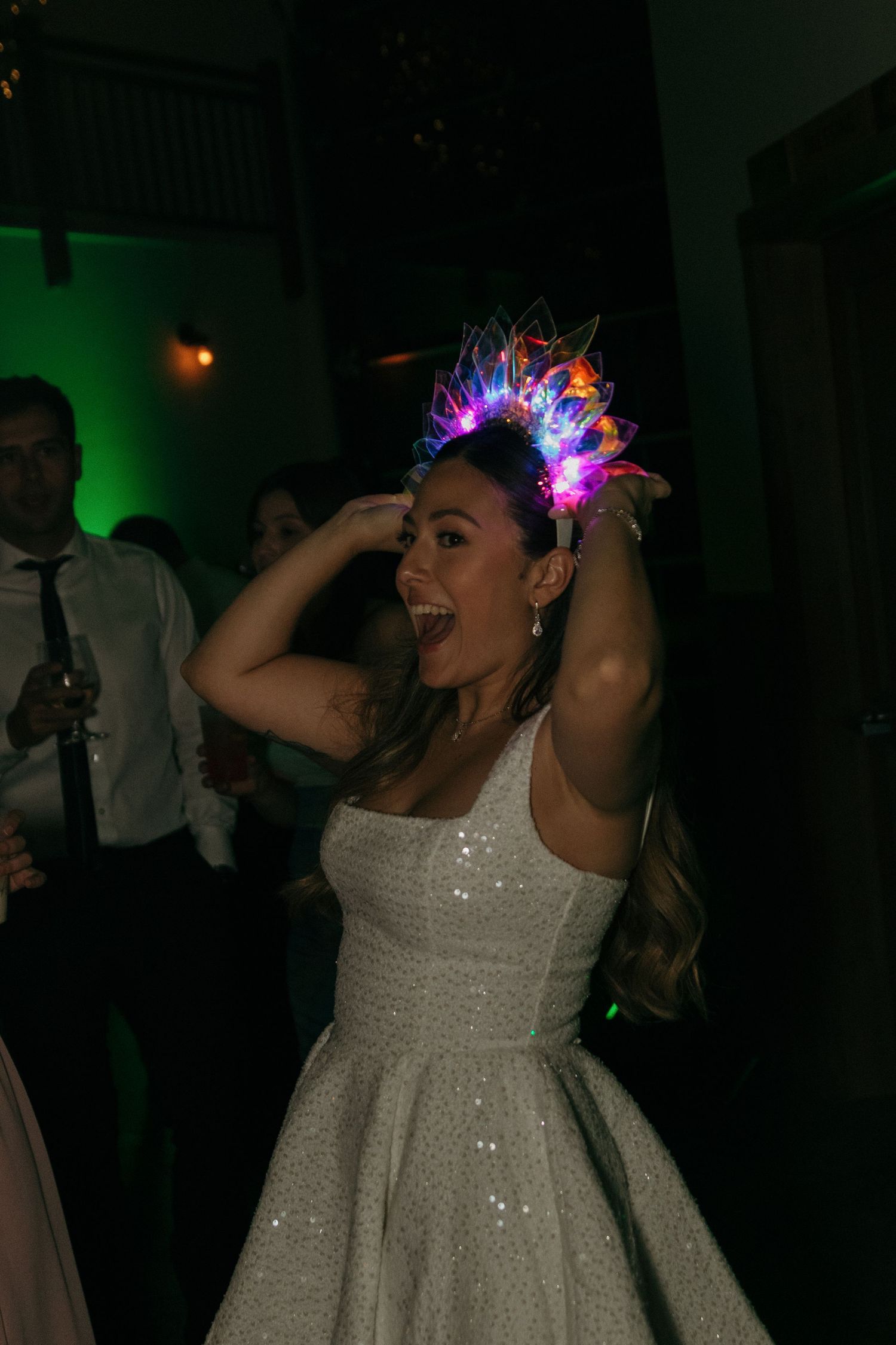 Wedding guests dance and celebrate on a dimly lit dance floor with colorful lighting effects.