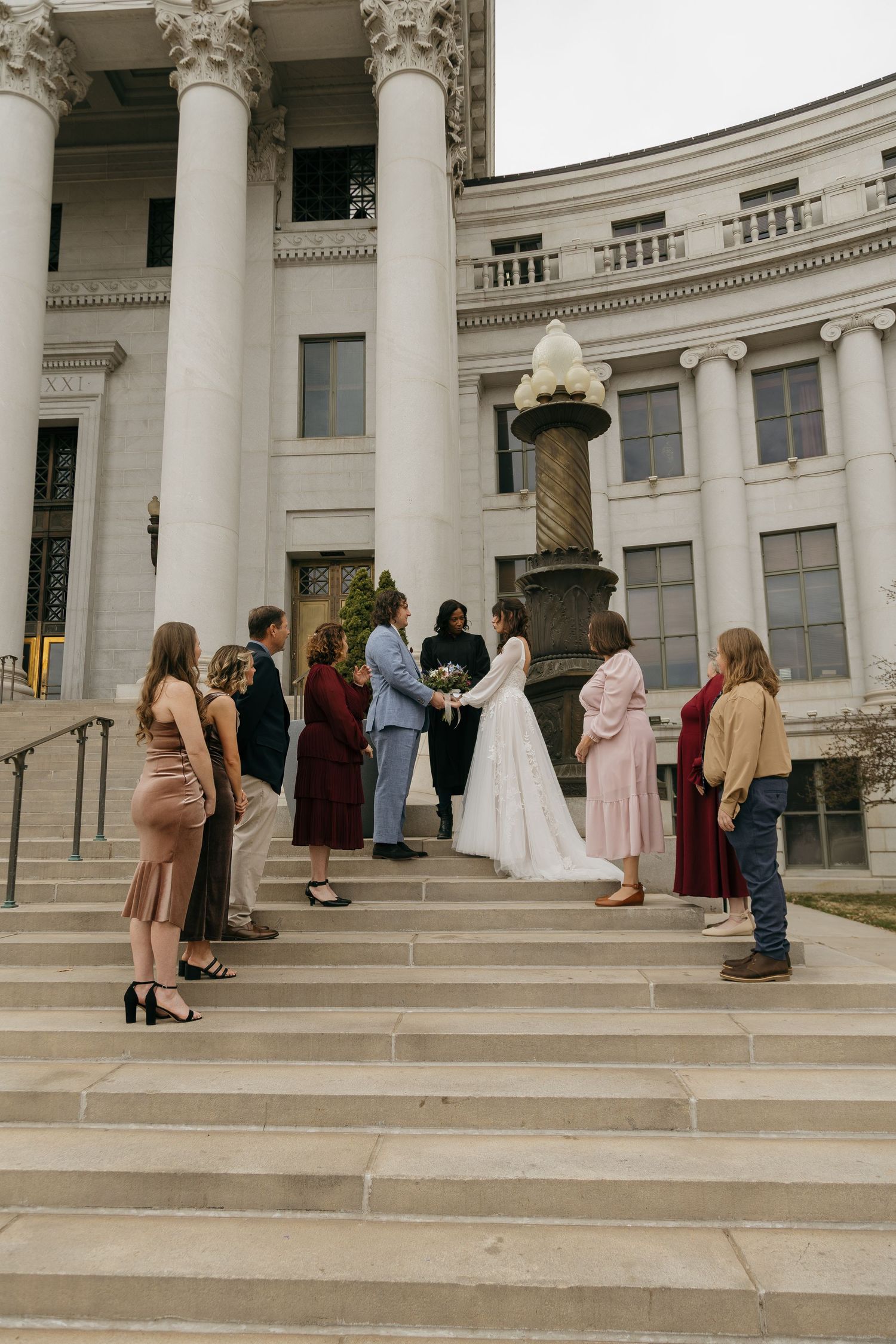 Wedding guests gather on grand stone steps of a neoclassical building with white columns.
