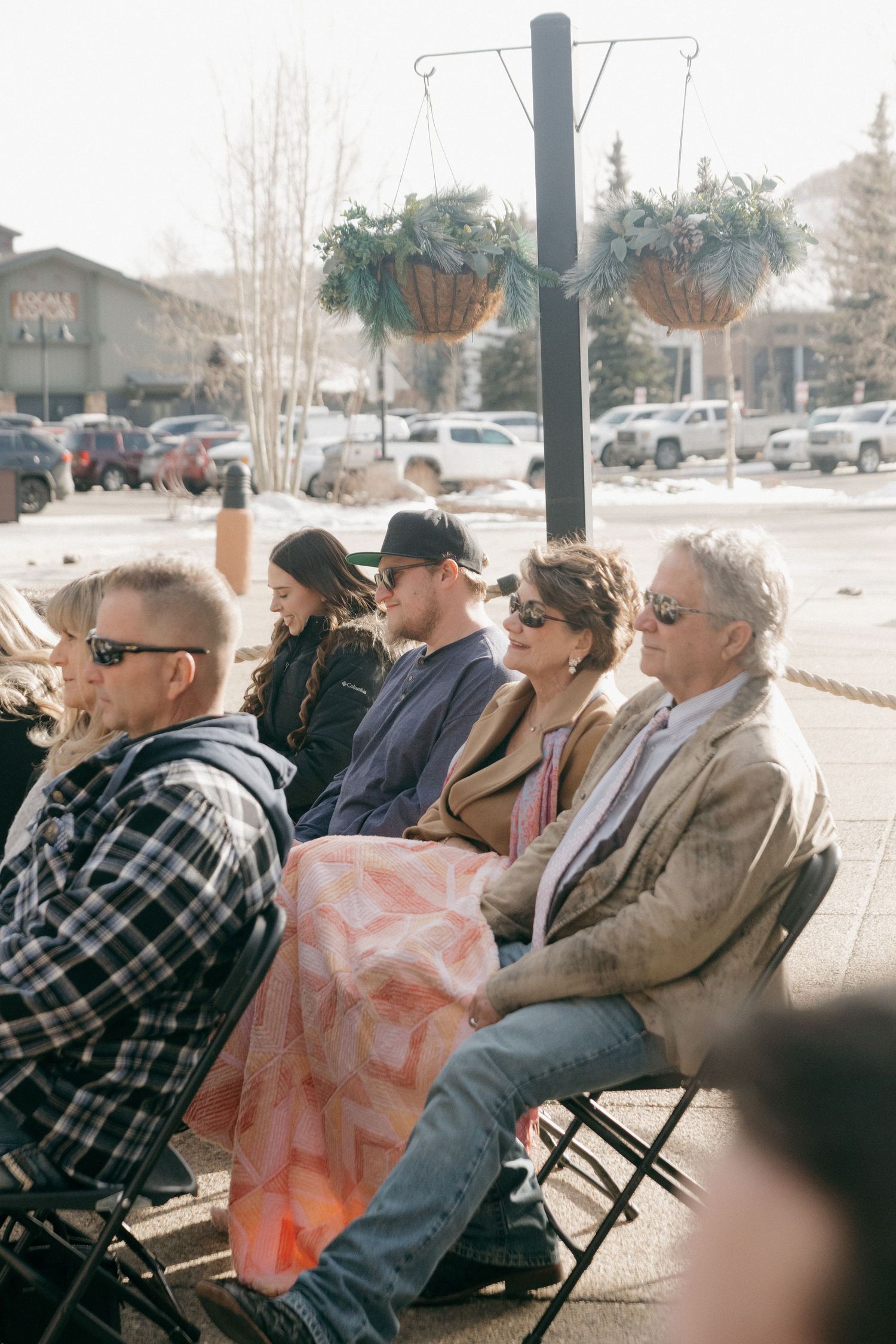 People sit in folding chairs outdoors at a community event with hanging planters and parked cars in the background.
