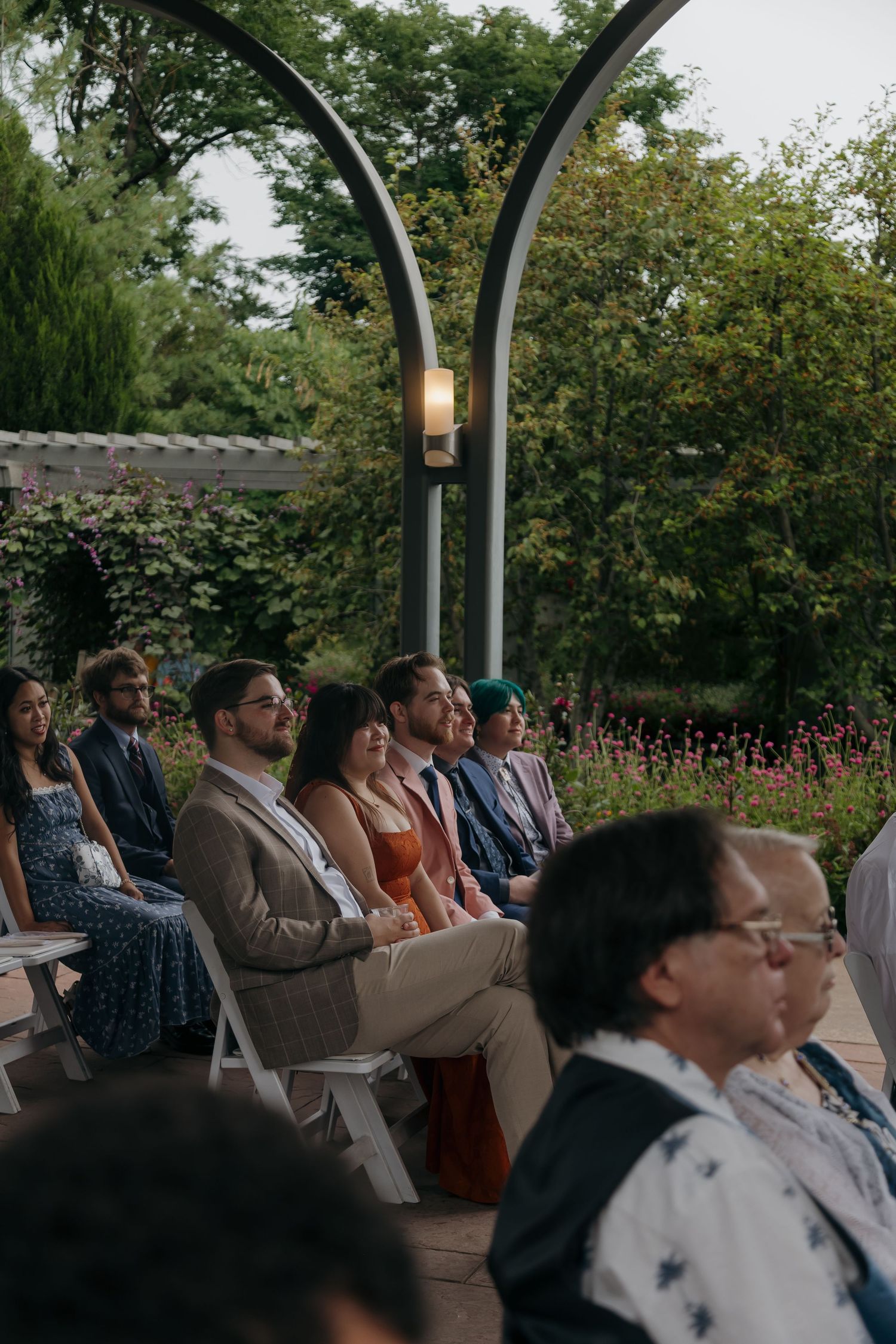 Wedding guests seated outdoors in garden ceremony under arched metal structure surrounded by lush greenery.