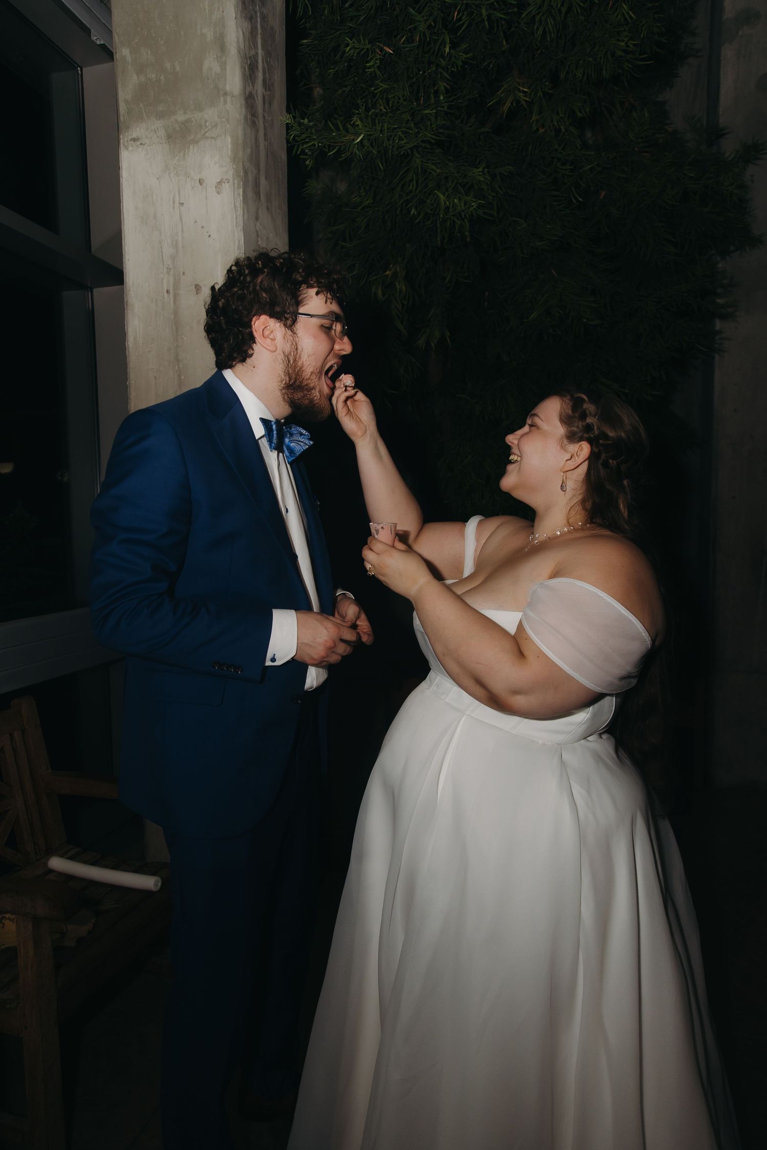 Bride feeds groom wedding cake while laughing together against dark green foliage backdrop at night.