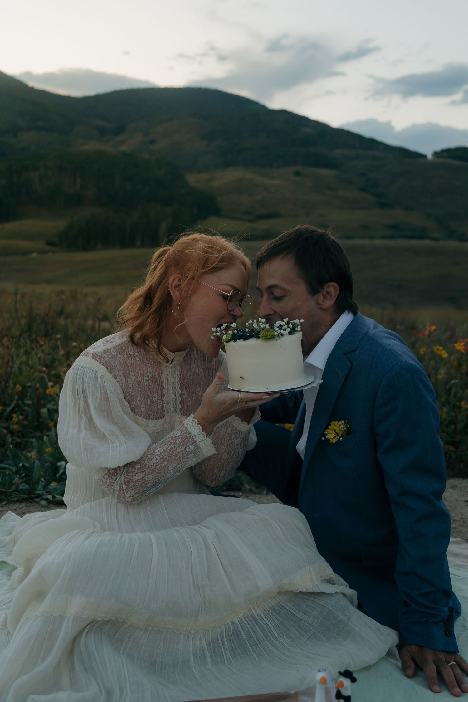 Bride and groom share an intimate moment with a white floral wedding cake outdoors against a scenic mountain backdrop.