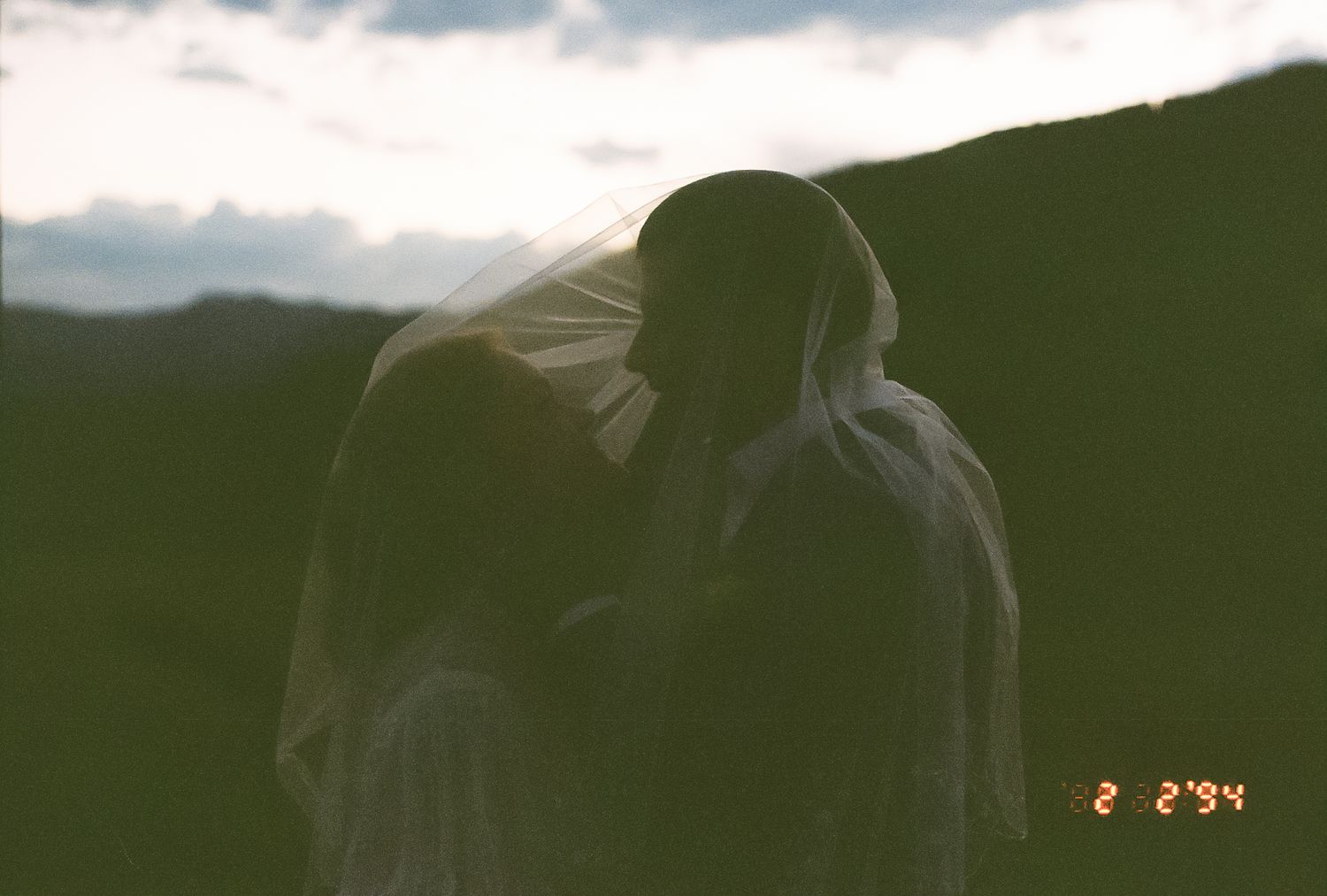 Silhouetted bride and groom share intimate kiss at dusk with dramatic cloudy sky background.