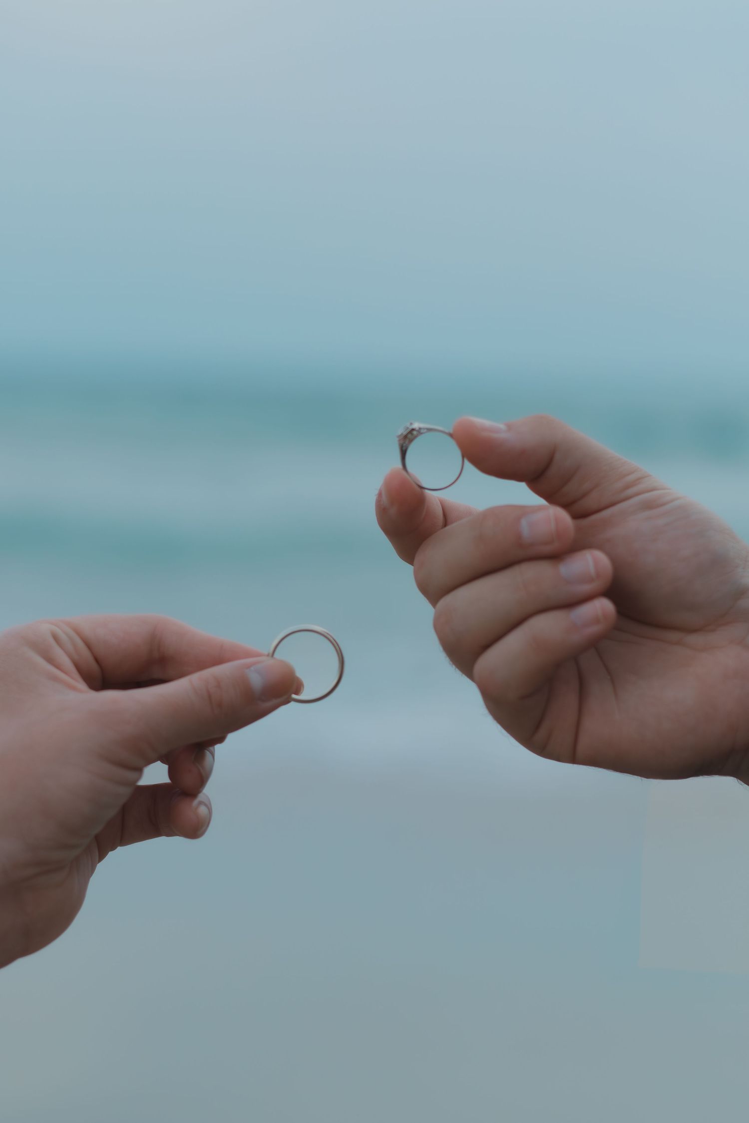 Two hands holding wedding rings near the ocean, symbolizing a romantic beach proposal or exchange of vows.