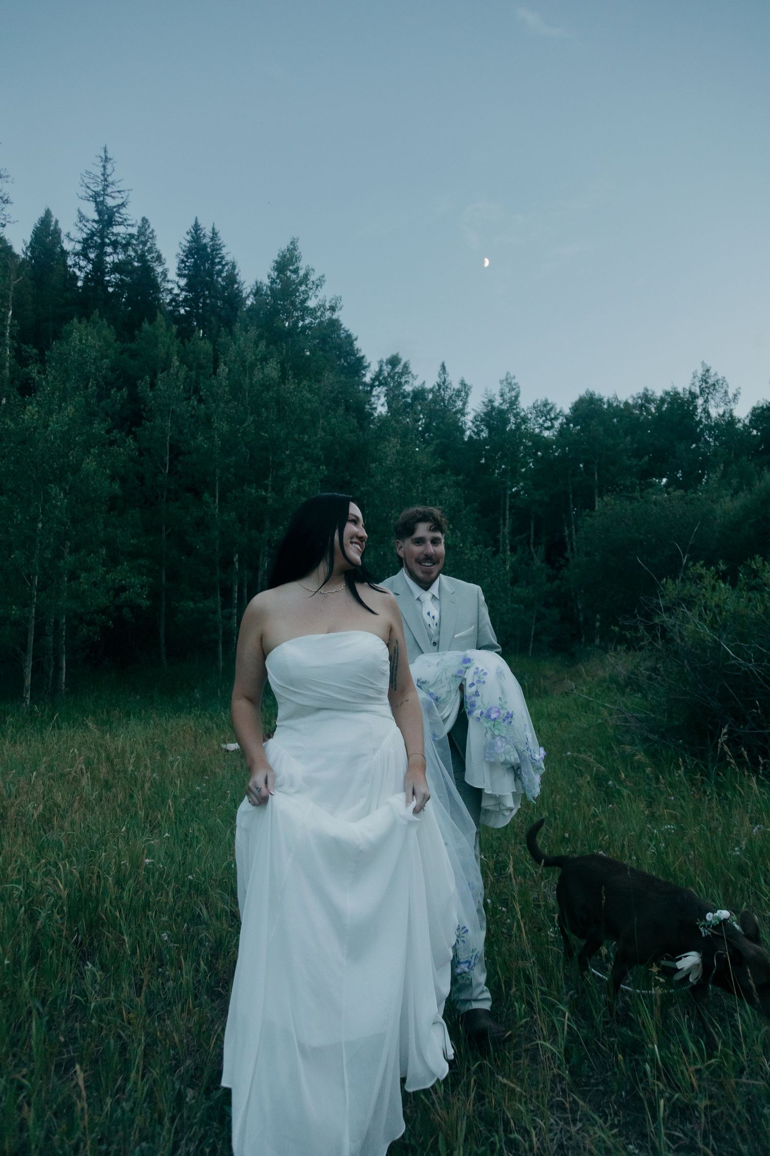 Laughing bride in white strapless gown walks with groom and dog through green meadow under crescent moon at dusk.