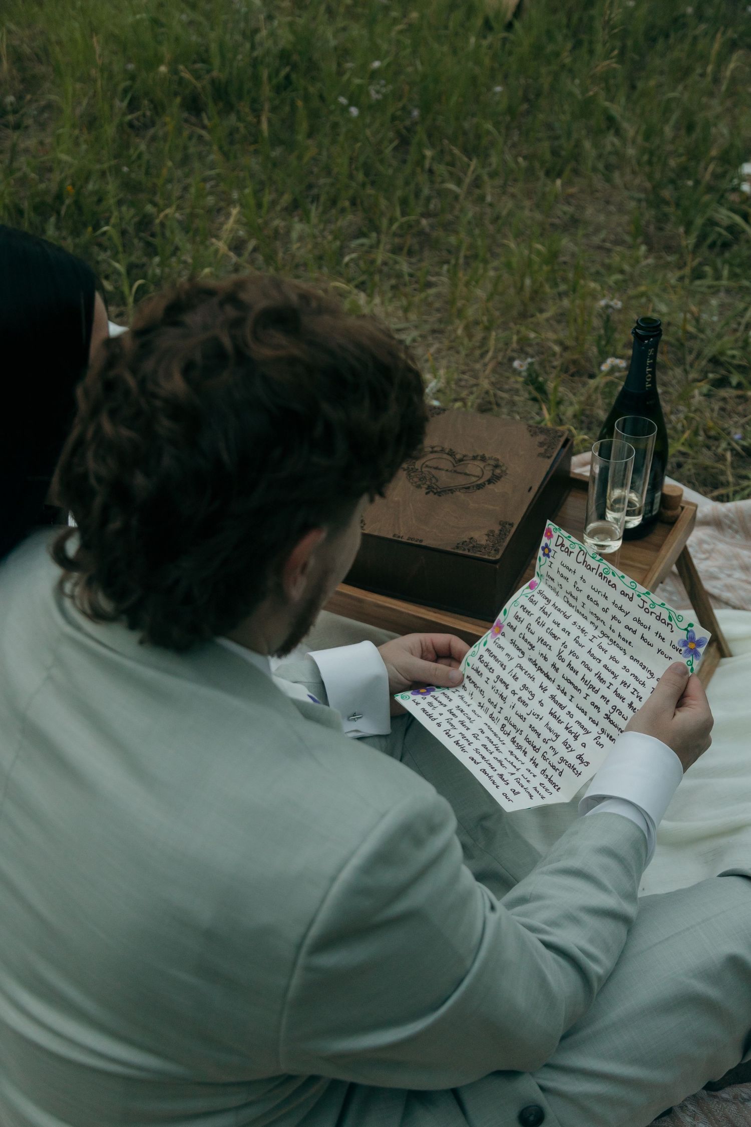 Groom in grey suit reads handwritten vows on a blanket outdoors with a bottle of wine and wooden box nearby.