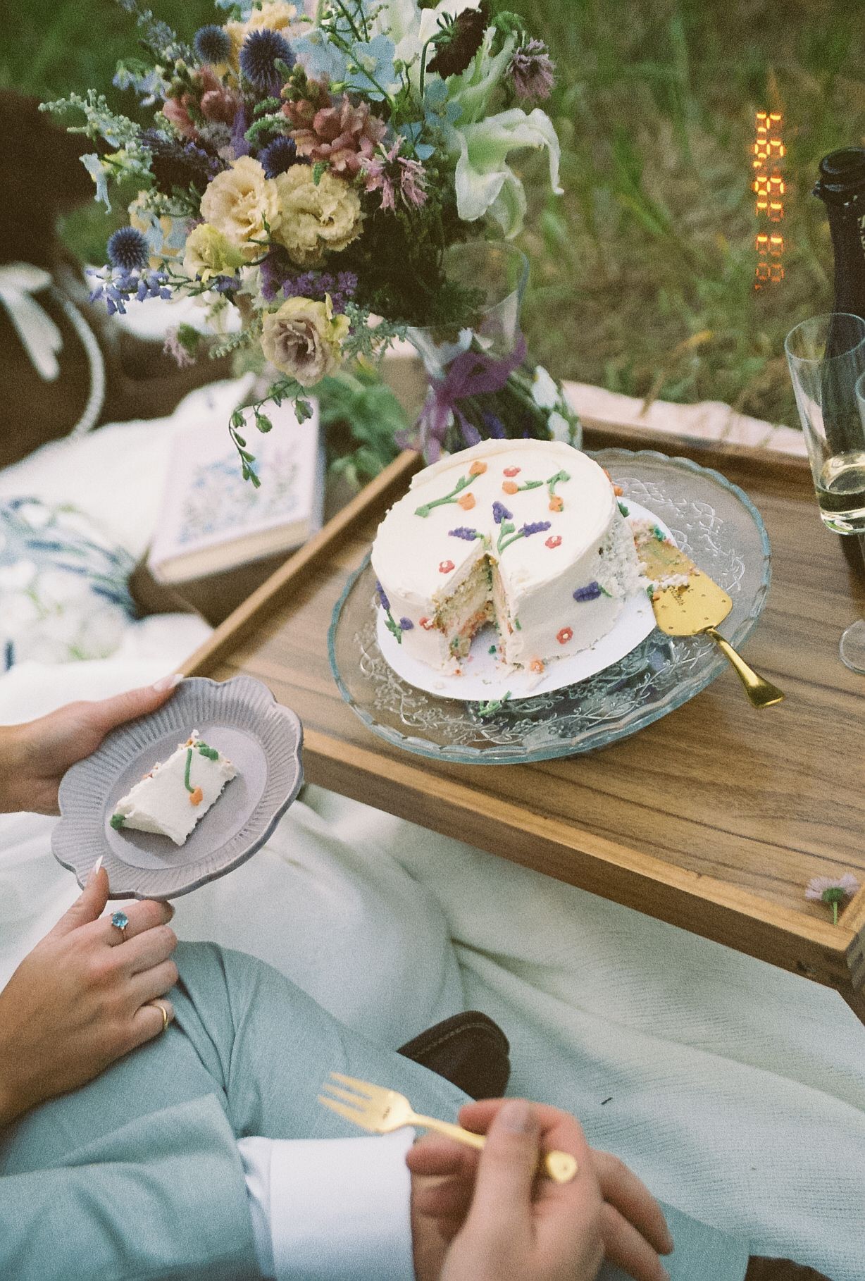 Couple enjoying confetti wedding cake on wooden picnic tray with wildflower bouquet during outdoor elopement.