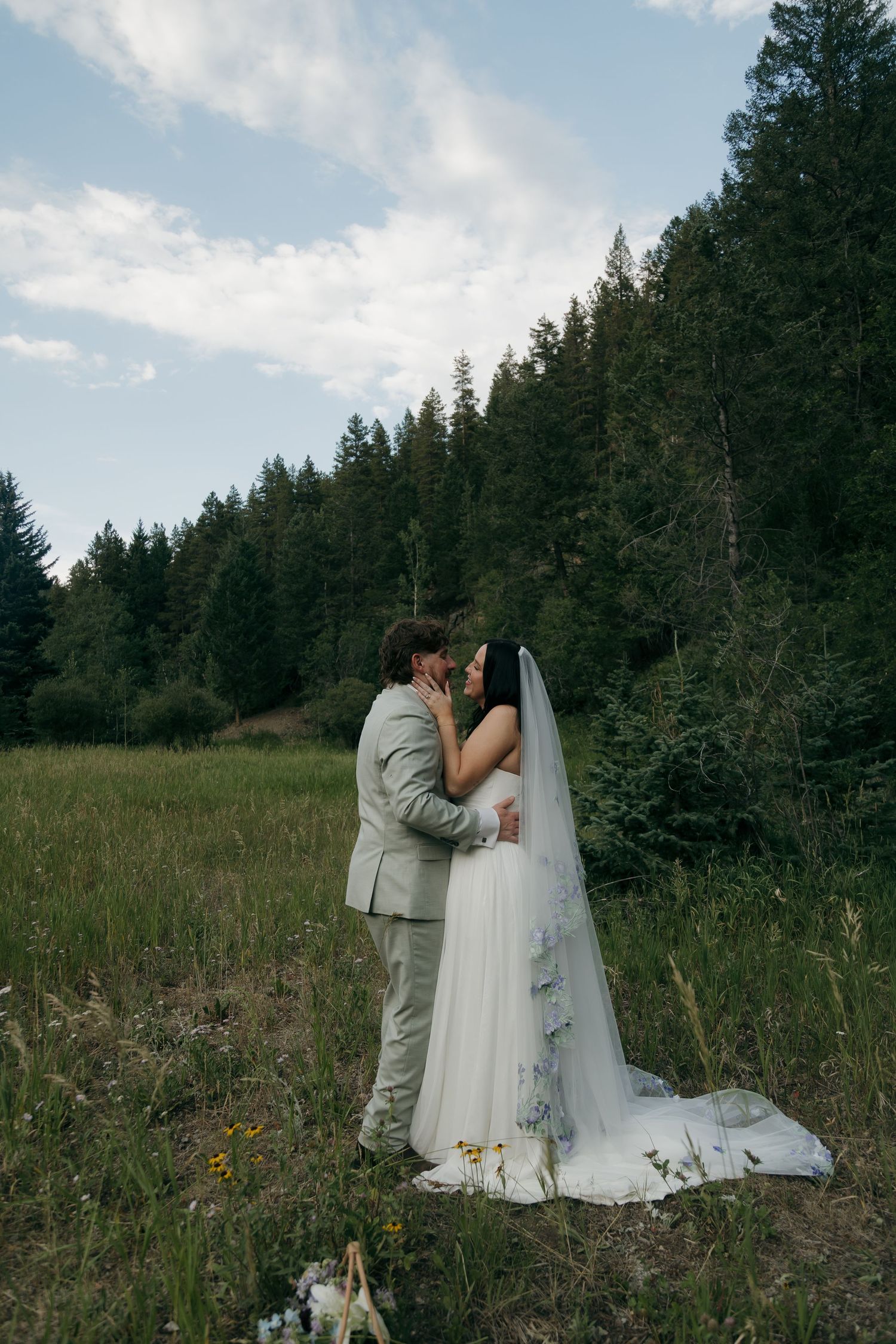 Bride and groom share a romantic kiss in a lush green meadow surrounded by tall pine trees on their wedding day.