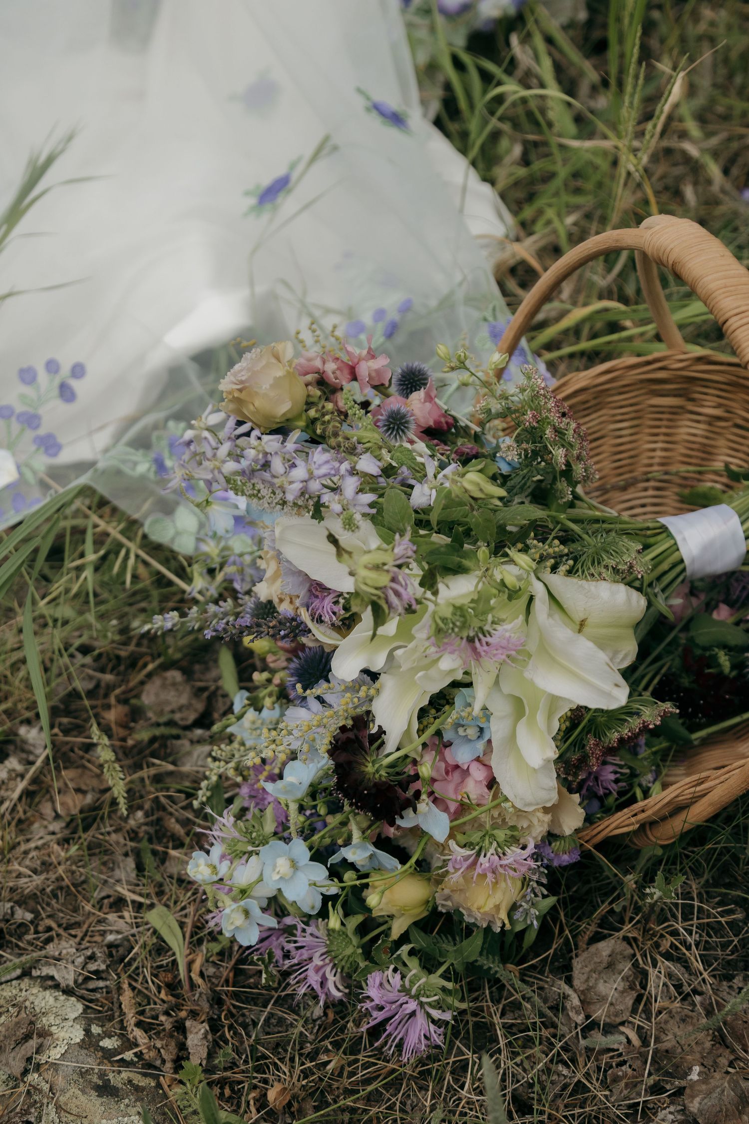 Wildflower bridal bouquet with white tulips, purple blooms beside wicker basket on forest ground.