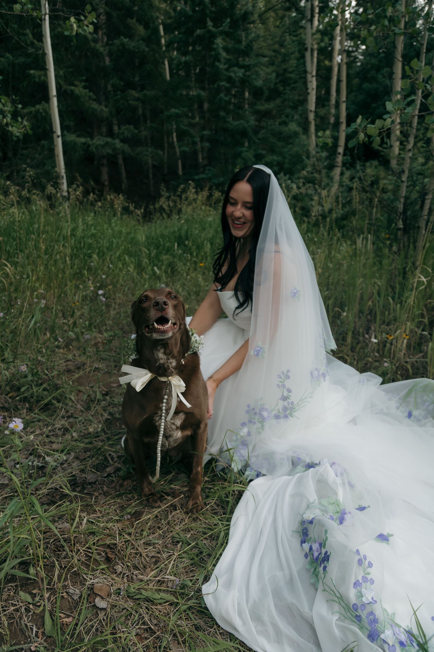 Laughing bride in floral veil kneels beside her brown dog wearing a bow in forest setting.