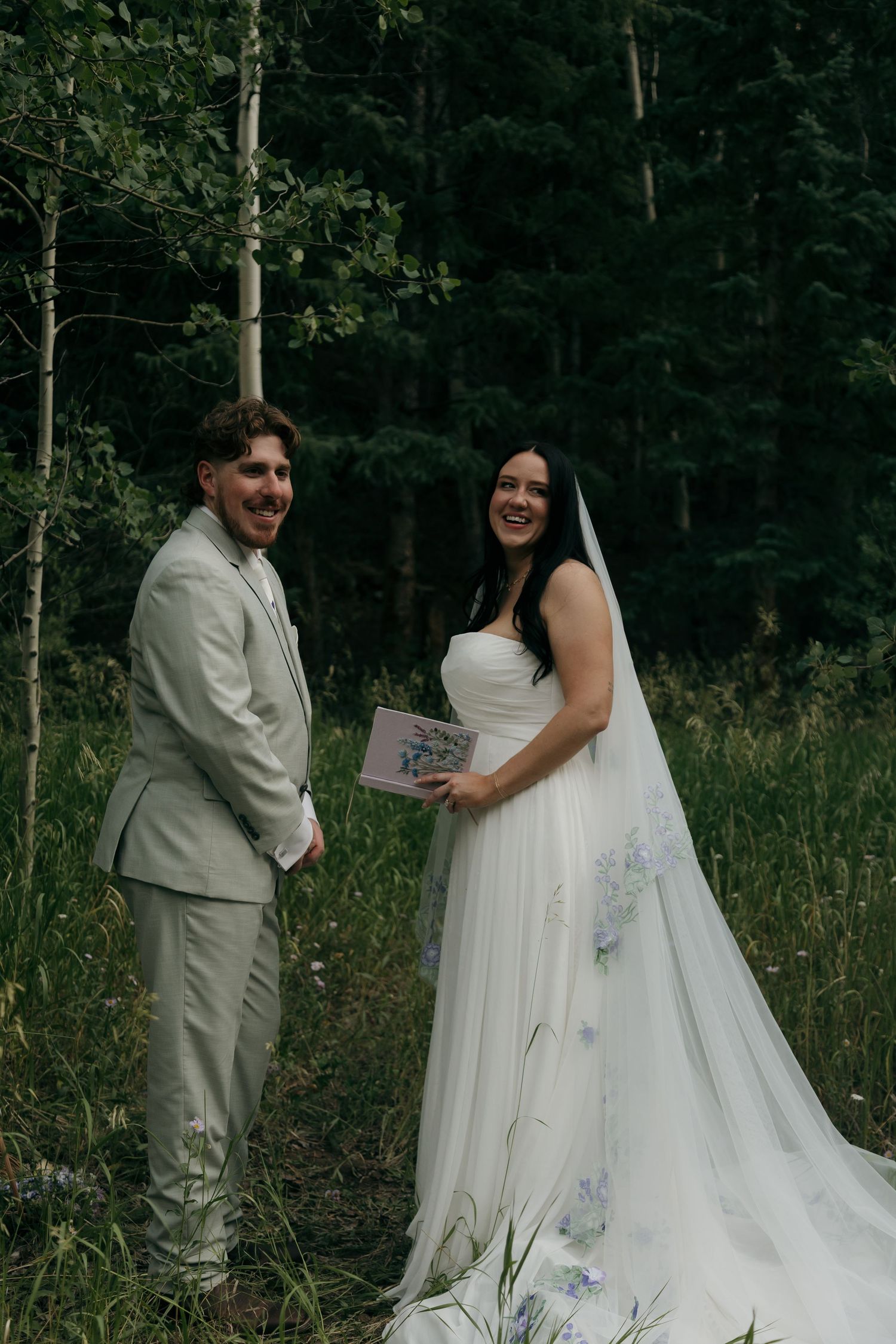 Laughing bride and groom pose together in a forest clearing, bride holding a gift box in her wedding attire.
