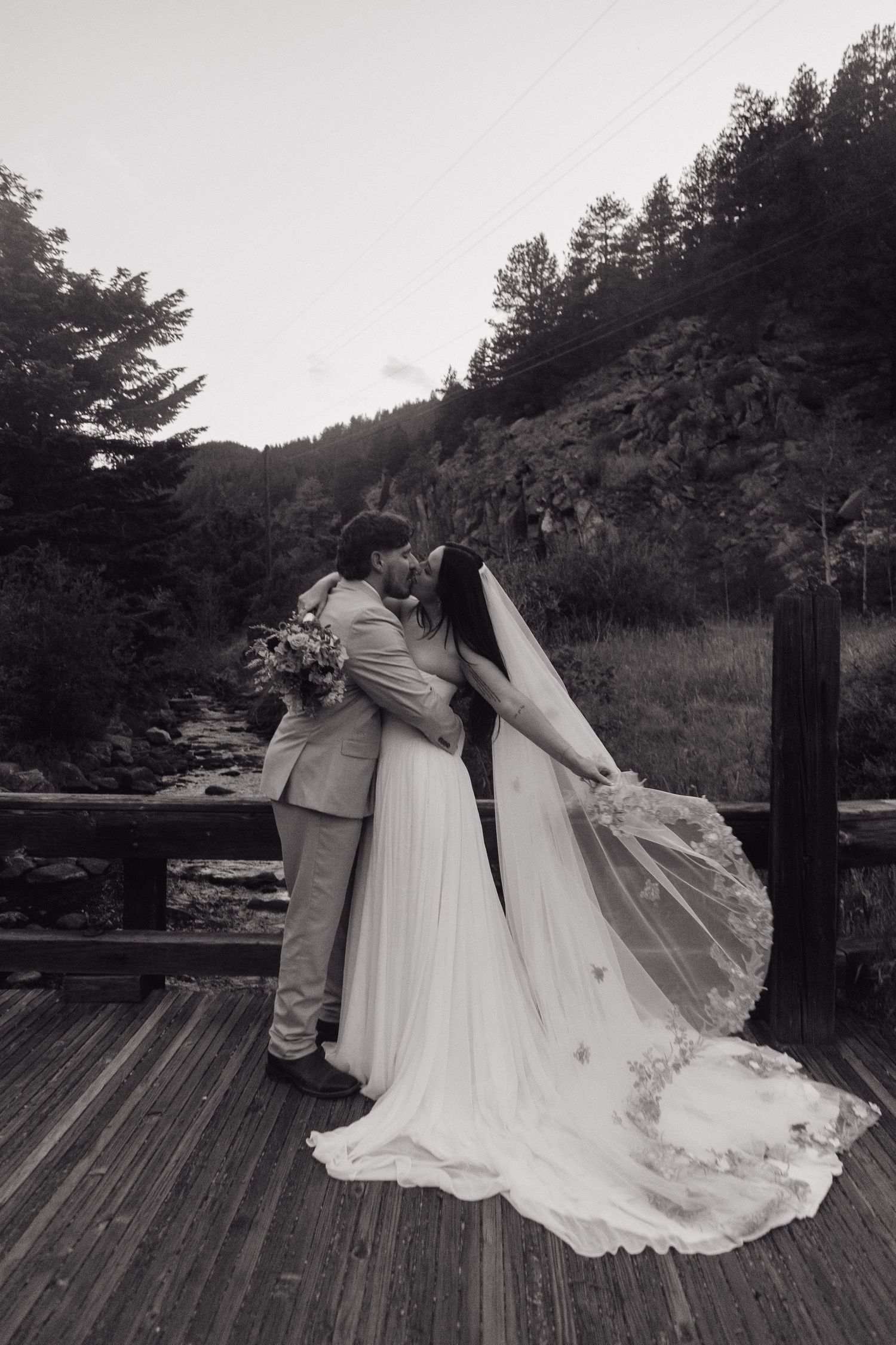Black and white photo of bride and groom sharing a romantic kiss on a wooden bridge, brides veil flowing in the wind.