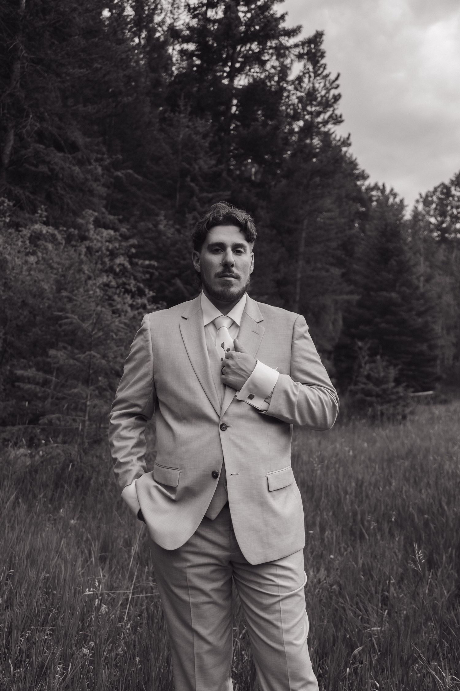 Groom in a light gray suit adjusts his tie while standing in a grassy field with evergreen trees behind him.