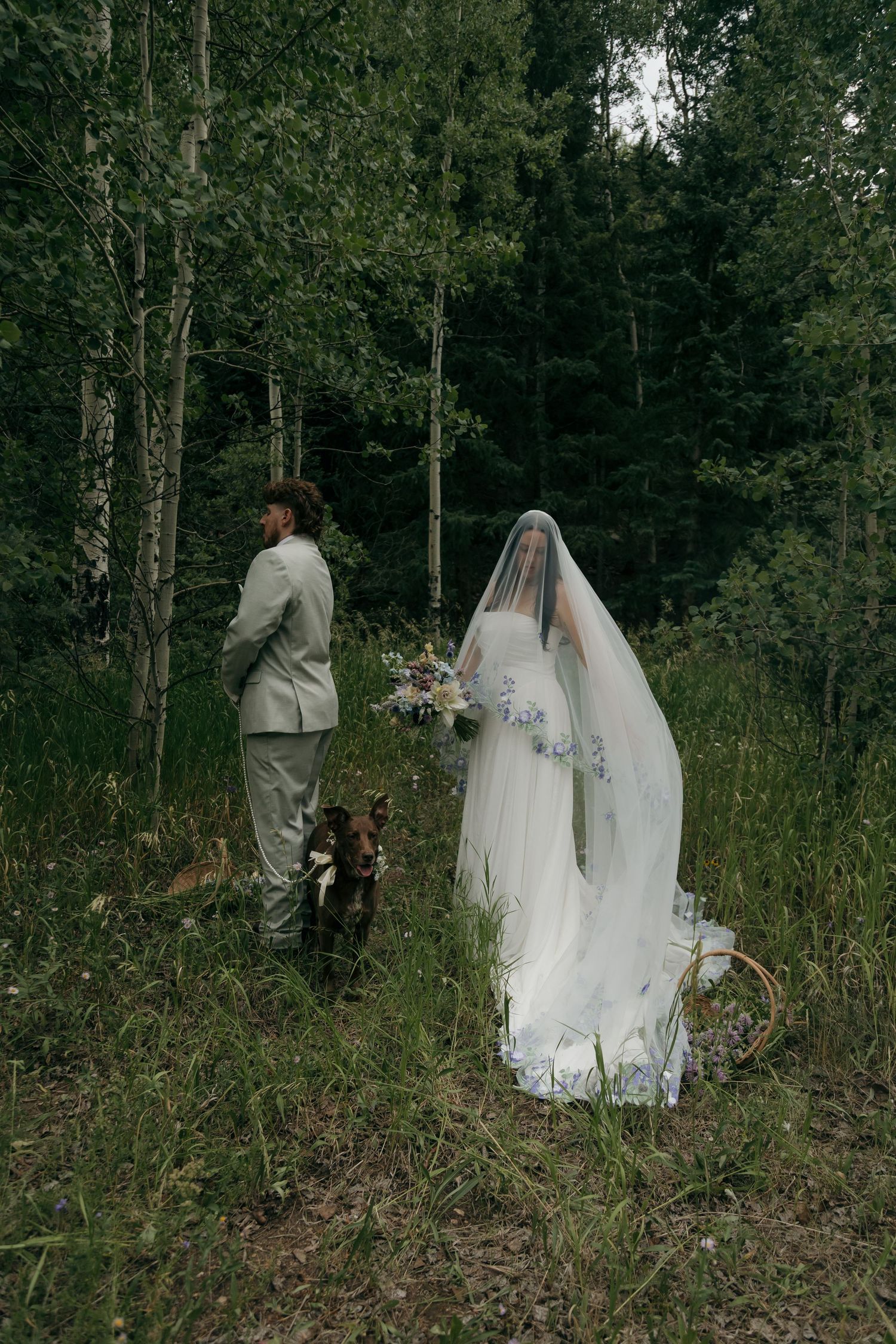 Bride in white gown and floral veil stands with groom in gray suit and dog in lush forest wedding photo.