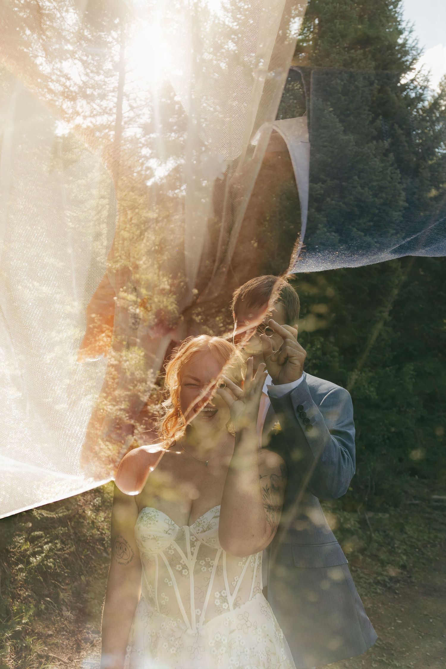 Bride and groom embrace beneath flowing veil with dreamy sunlit forest bokeh effect.