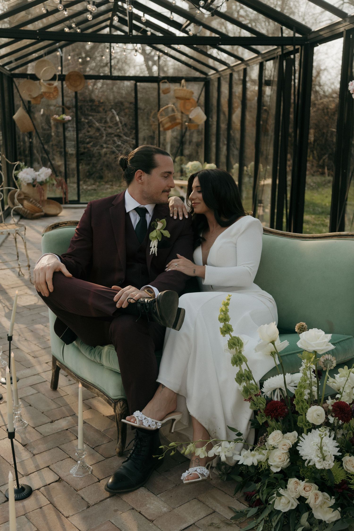 A wedding couple sits intimately on a vintage couch inside a greenhouse decorated with white flowers and romantic decor.