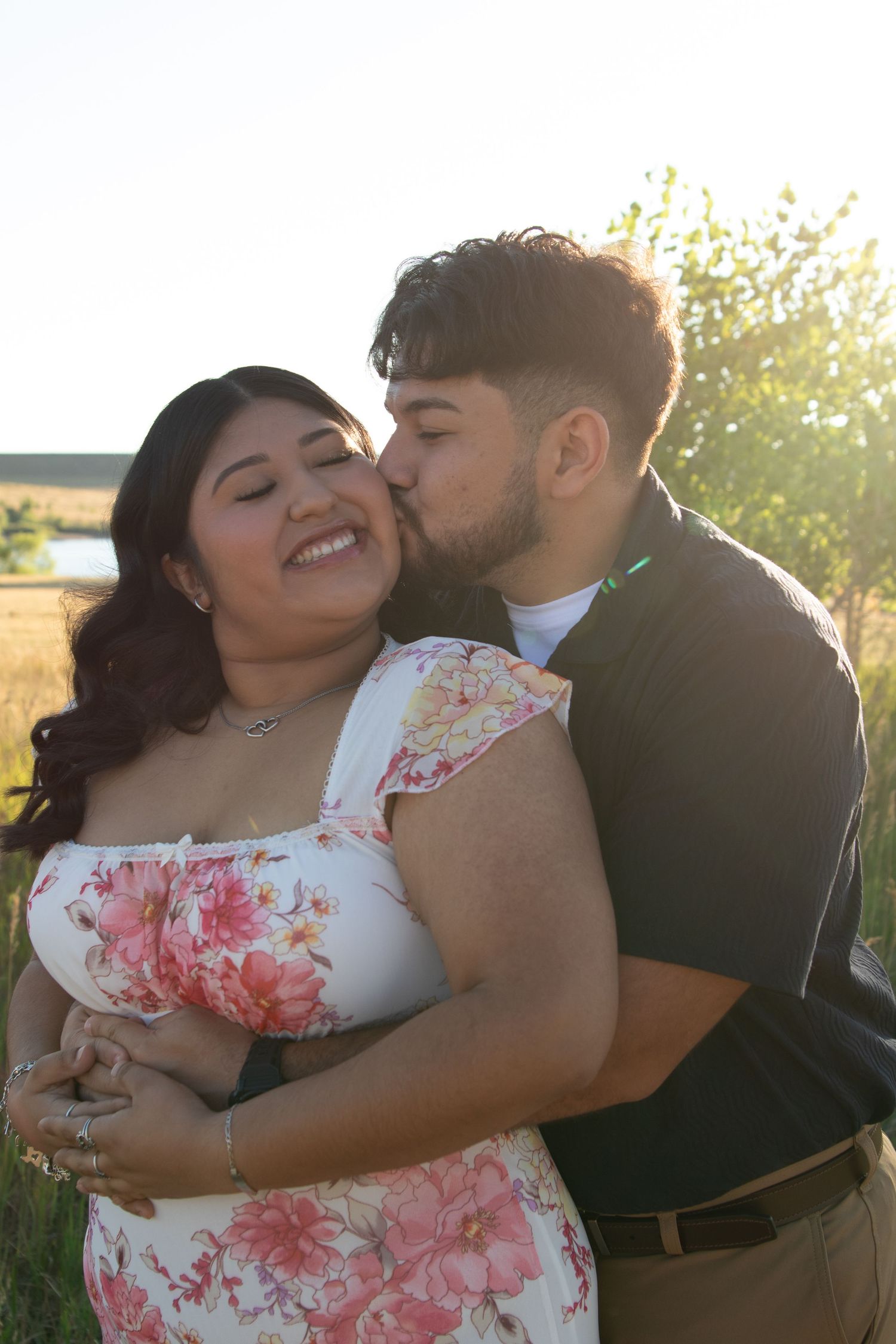 A romantic couple embraces in a sunlit field, with her wearing a floral white dress and him in dark clothing.