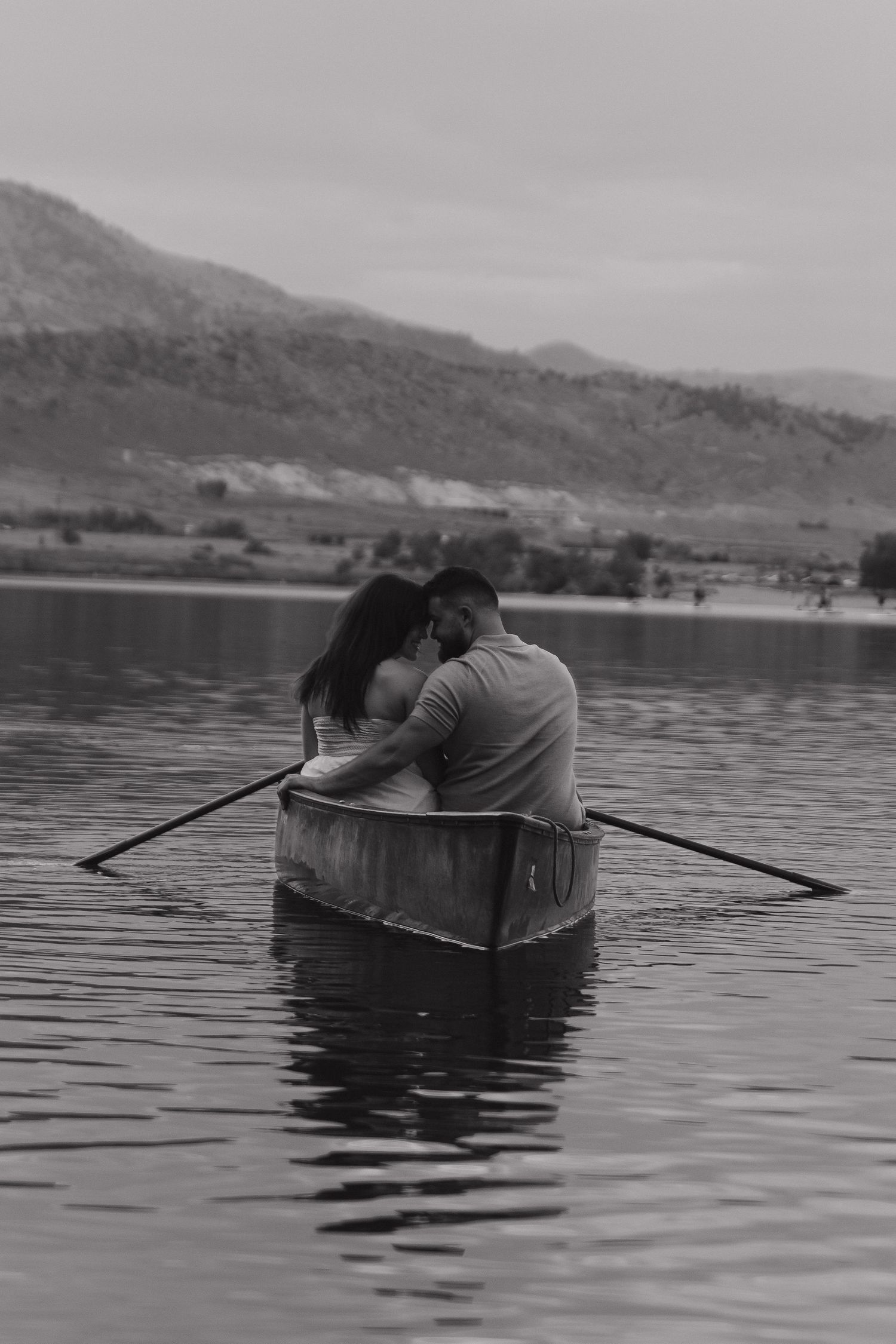 A couple embraces while rowing a canoe on a peaceful lake with mountains in the background.