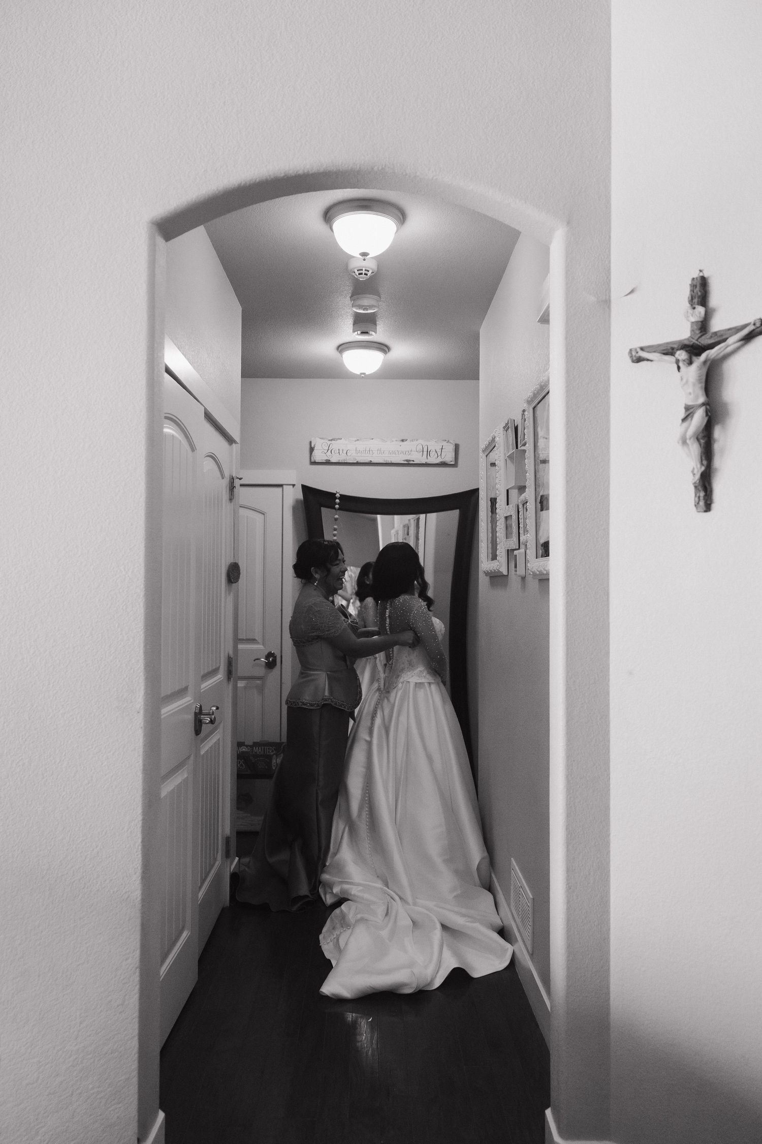 Black and white photo of a bride being helped into her wedding gown in a hallway with a crucifix on the wall.