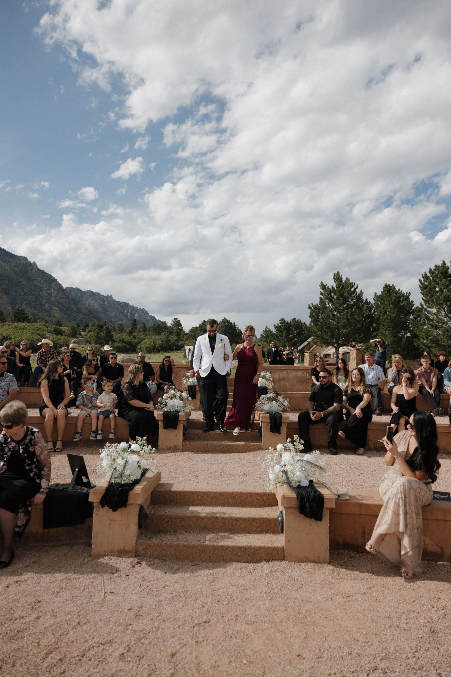 Outdoor wedding ceremony with guests seated on wooden benches under blue sky with mountain backdrop.