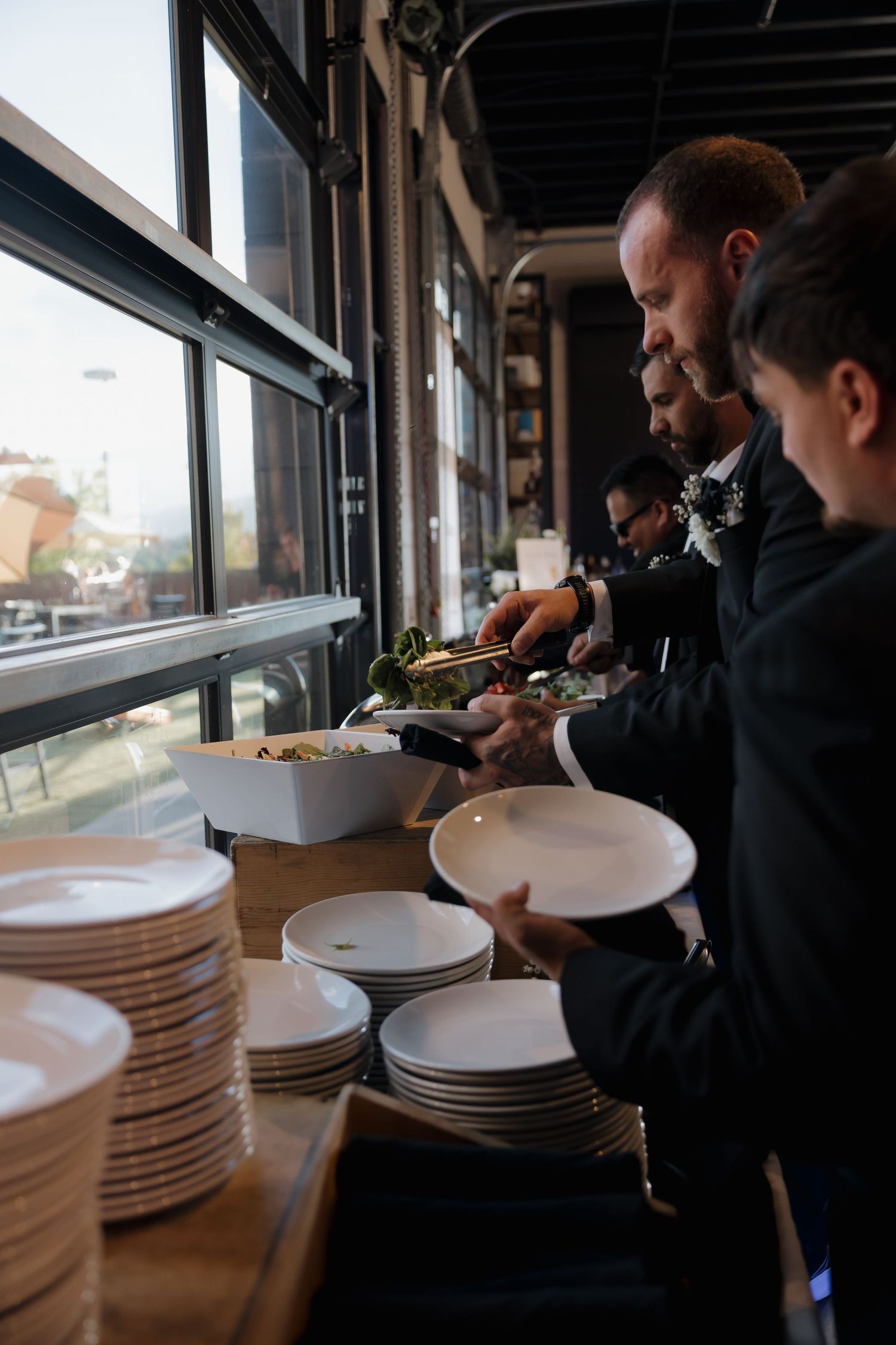 Groomsmen in black suits serve themselves at wedding reception buffet table by large windows.