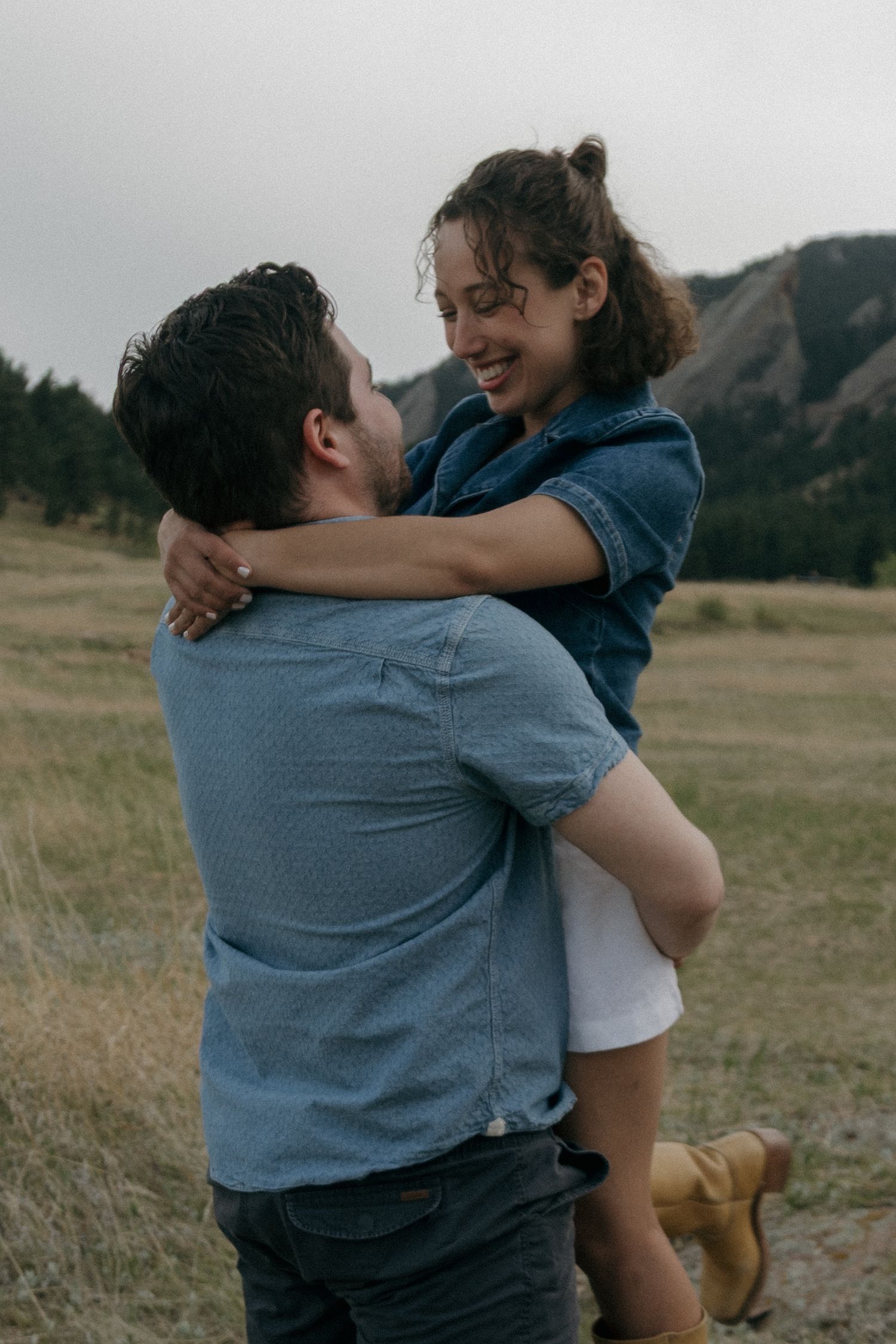 A joyful couple embraces in a mountain meadow, the man lifting the smiling woman in a denim shirt outdoors.