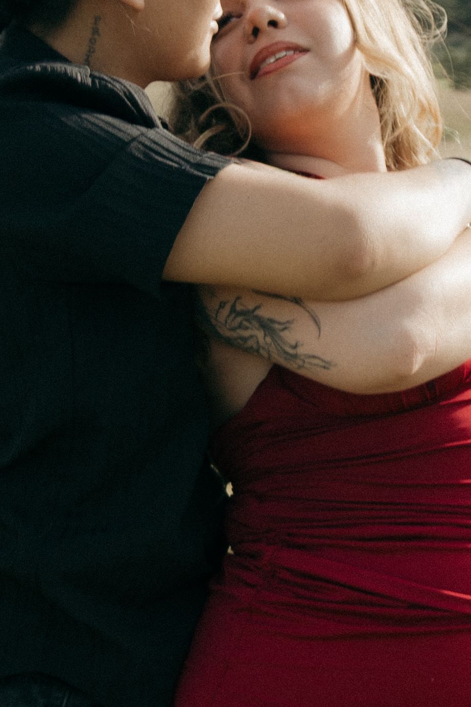 A couple embraces intimately, woman in red dress with tattoos wrapping arms around partner in black shirt.