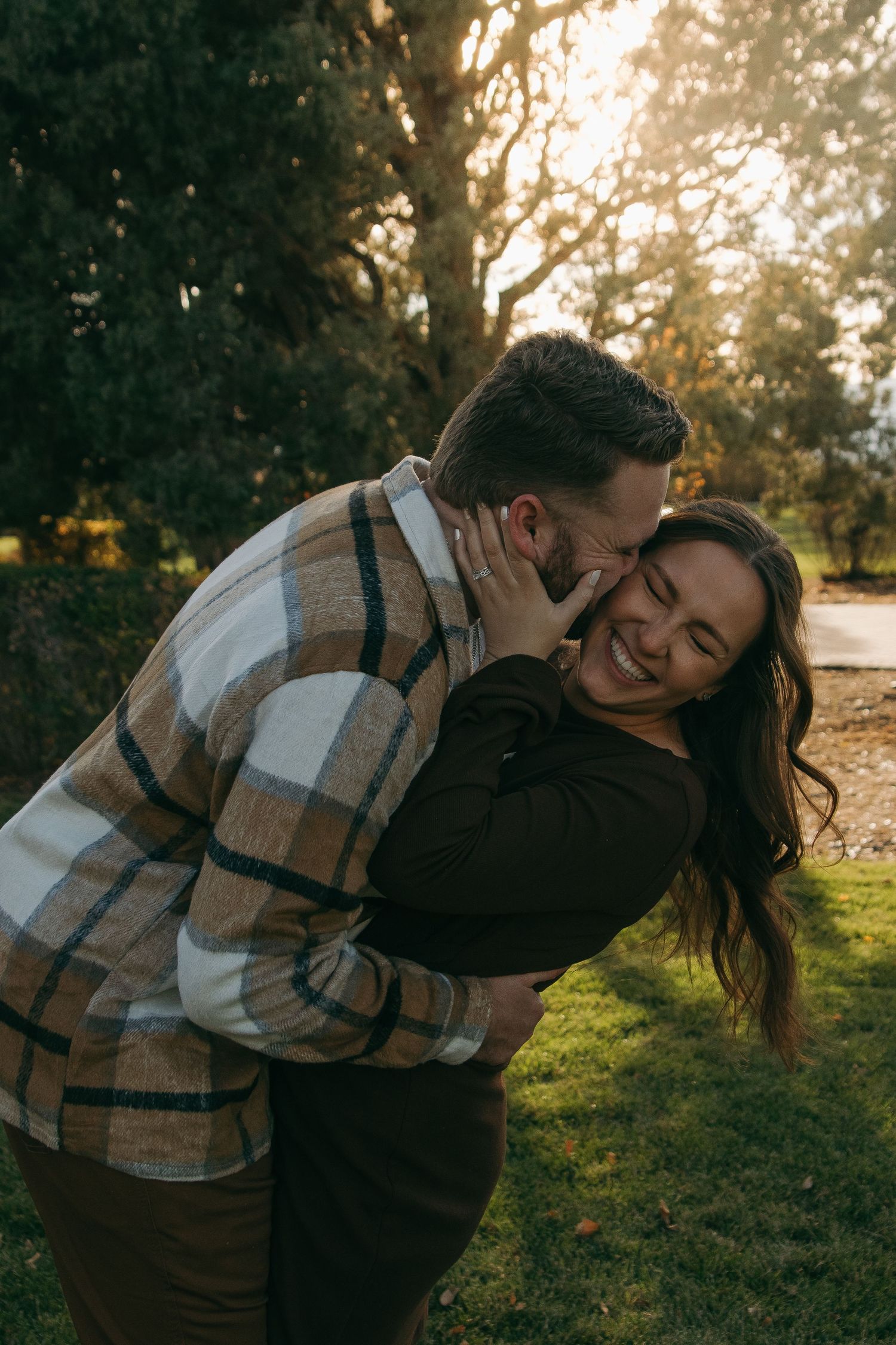 Couple embracing outdoors at golden hour, man kissing woman's cheek as she laughs joyfully in a park setting.