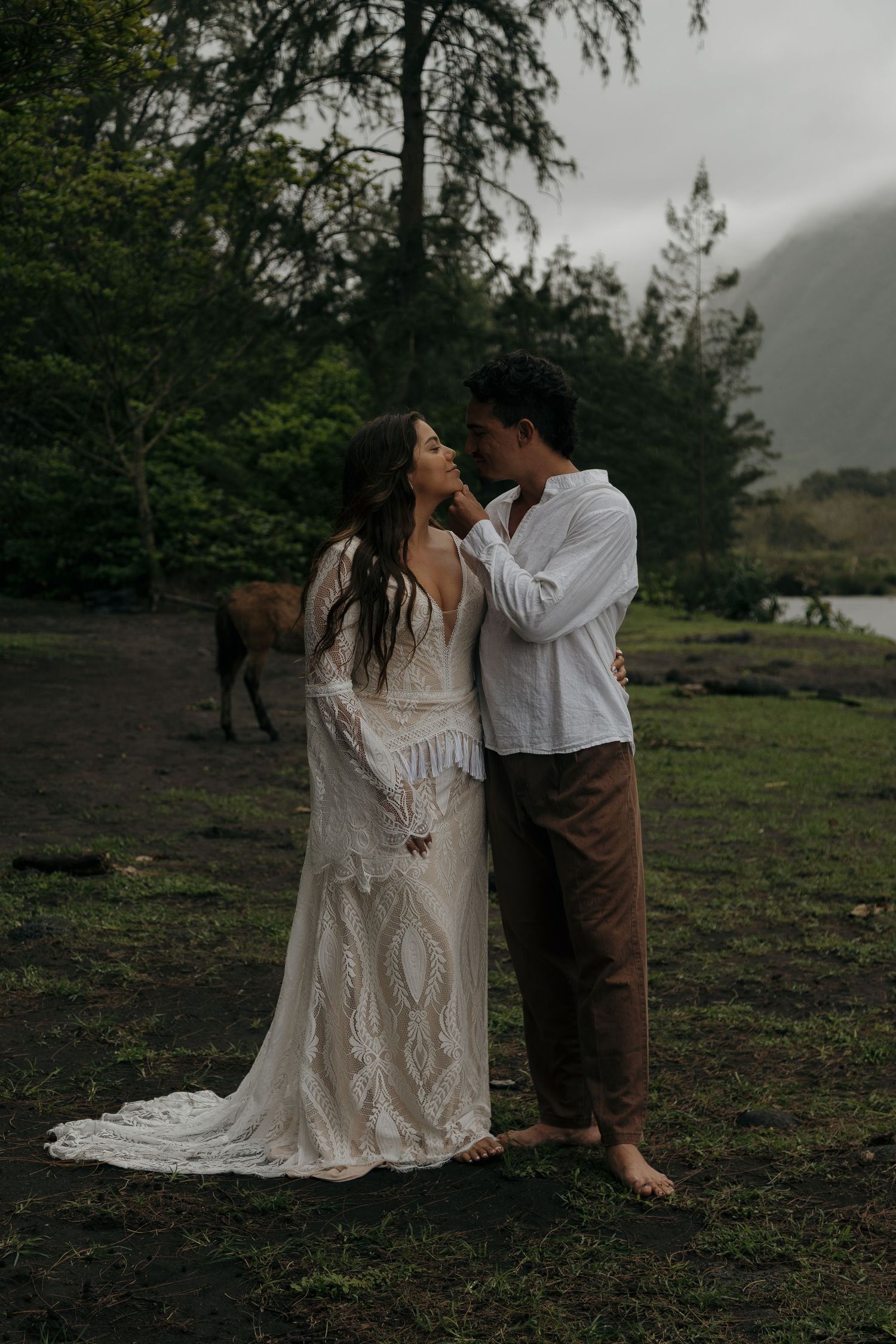 Couple in bohemian wedding attire share a romantic moment in a lush forest with a horse grazing nearby.
