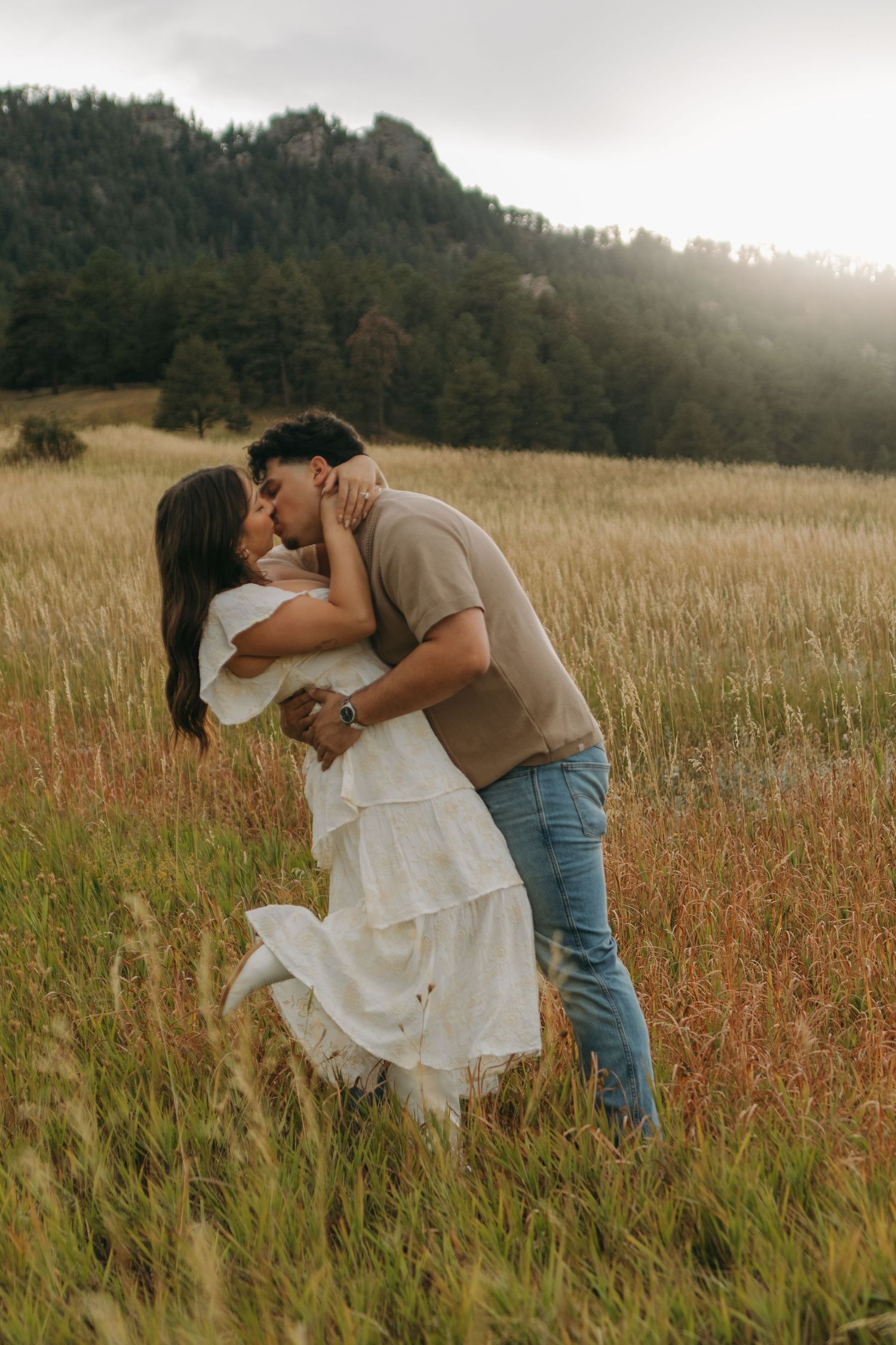 Couple sharing romantic dip kiss in tall golden grass field with mountain backdrop.