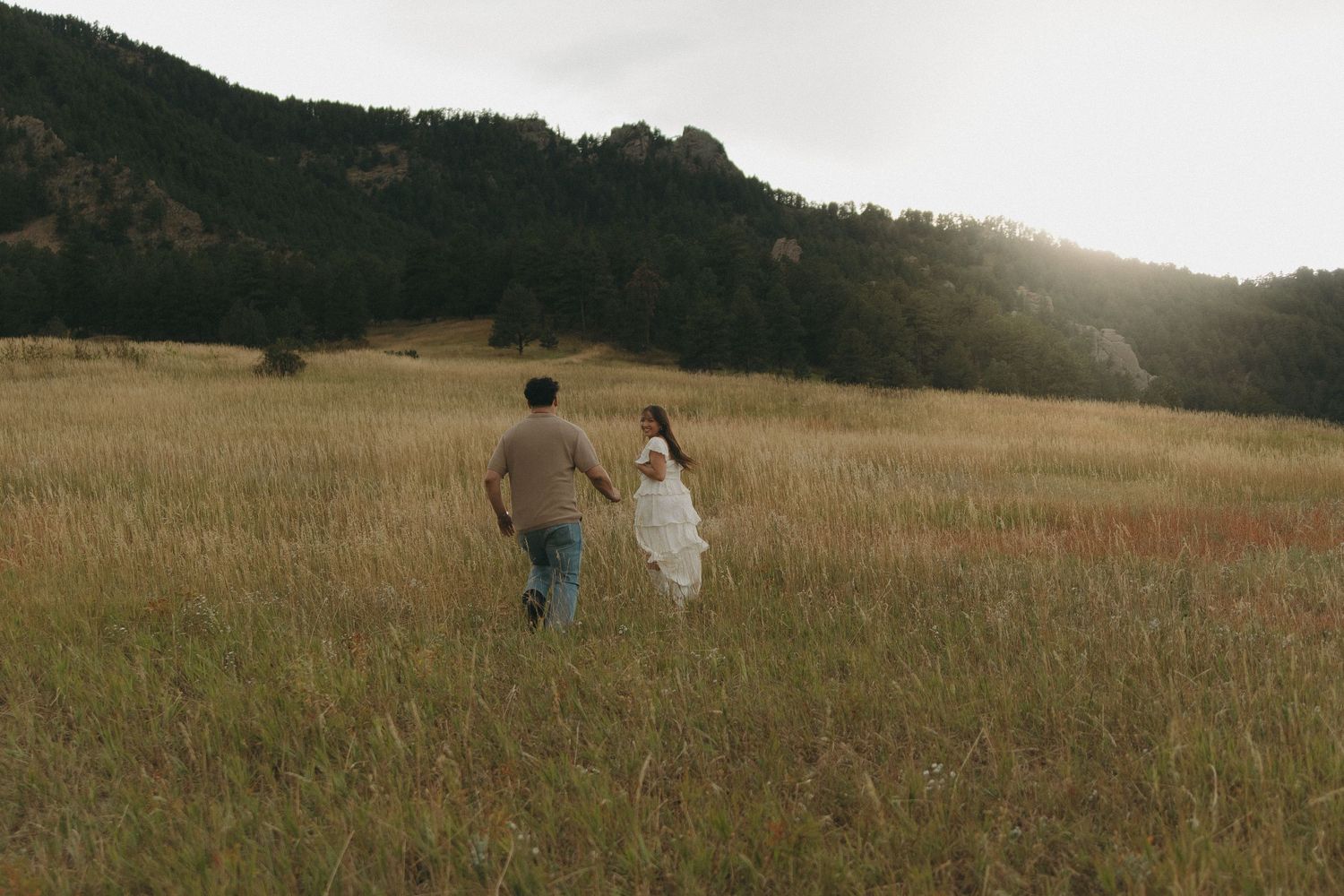 Couple holding hands running through golden meadow with mountain backdrop at sunset.