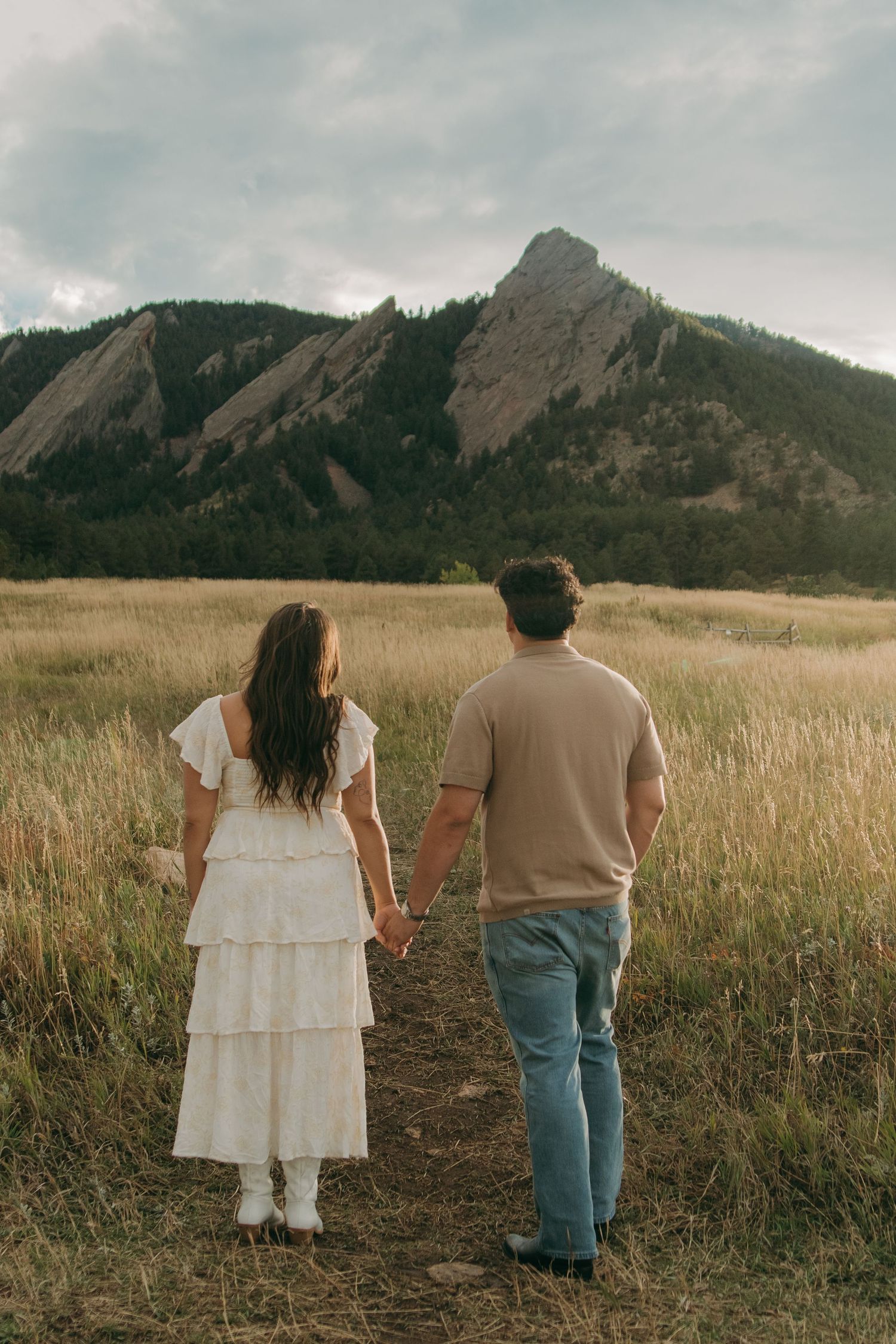 Couple holding hands walking toward the dramatic Flatirons rock formations under a cloudy Colorado sky.