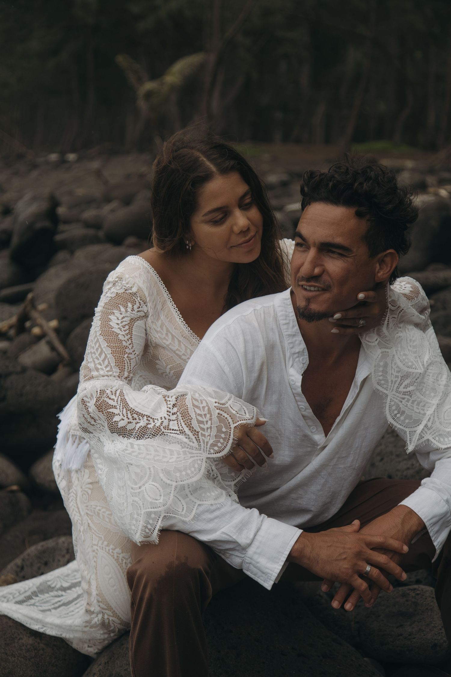 Couple sitting on coastal rocks, bride in boho lace gown resting hand on grooms shoulder.