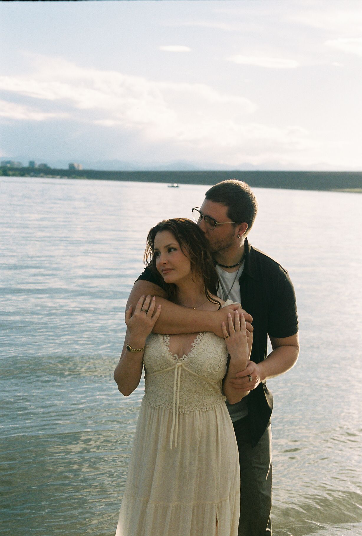 Couple embracing by a lake at sunset, woman in white lace dress, man in black shirt holding her shoulders.