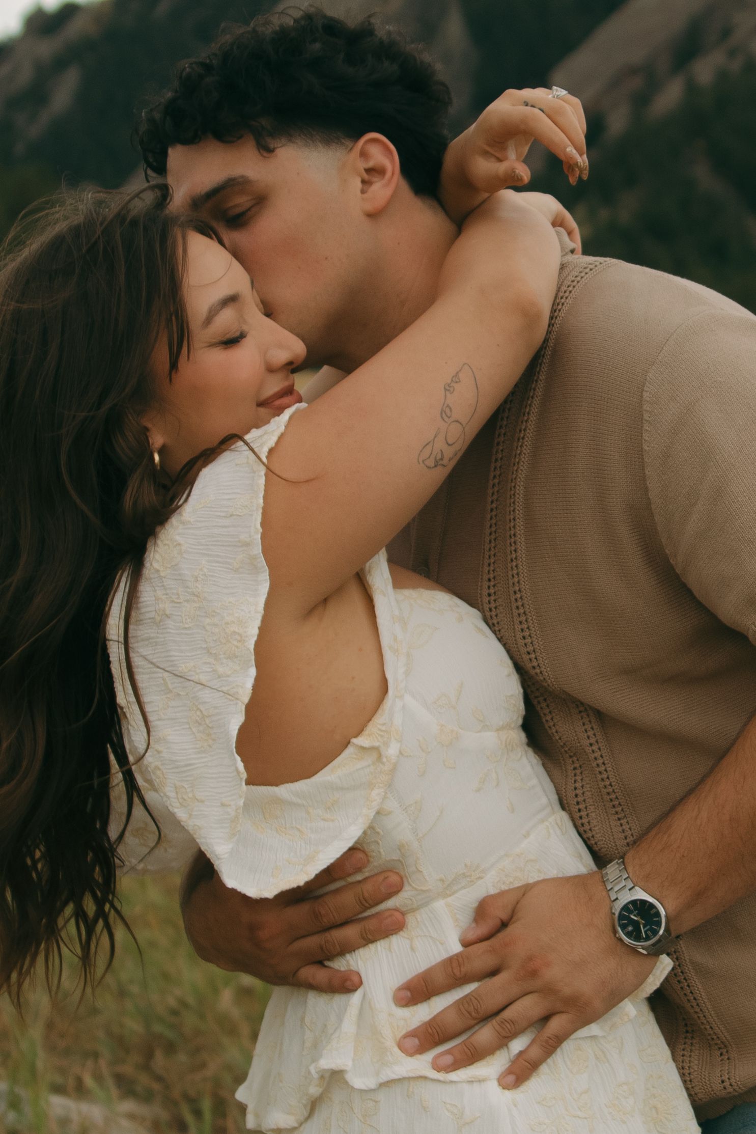 A couple shares an intimate embrace with the man kissing the woman's cheek in a mountain setting.