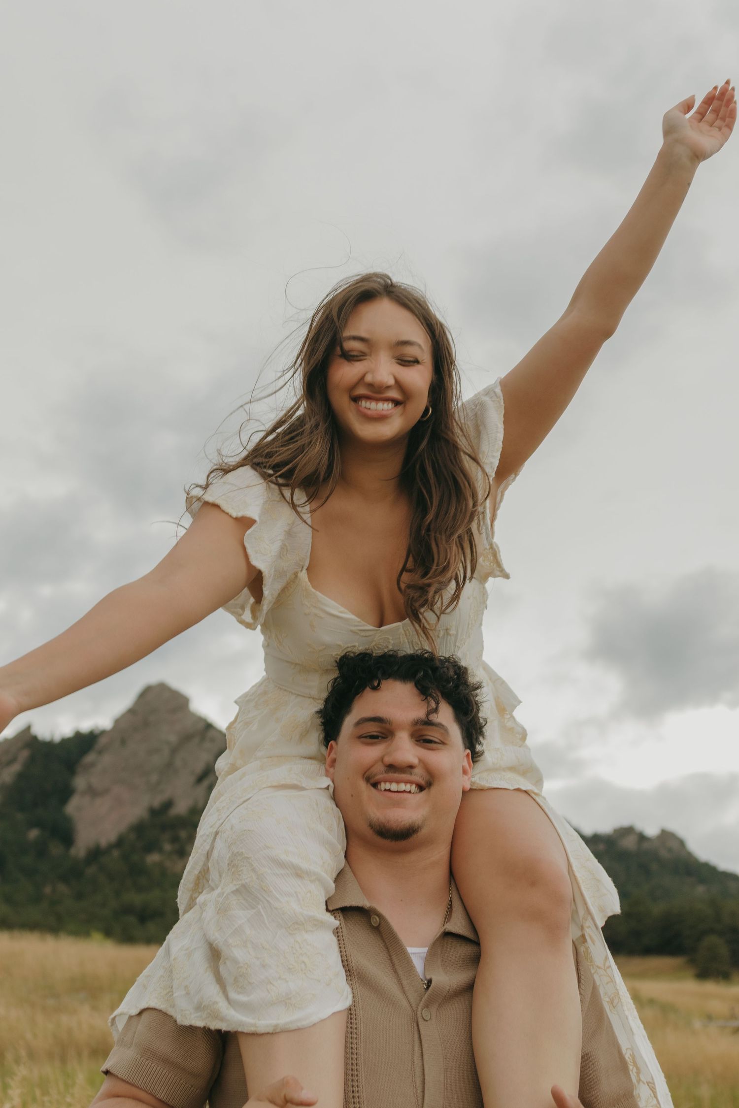 Joyful woman sitting on mans shoulders with arms raised in field with rocky mountain flatirons backdrop.