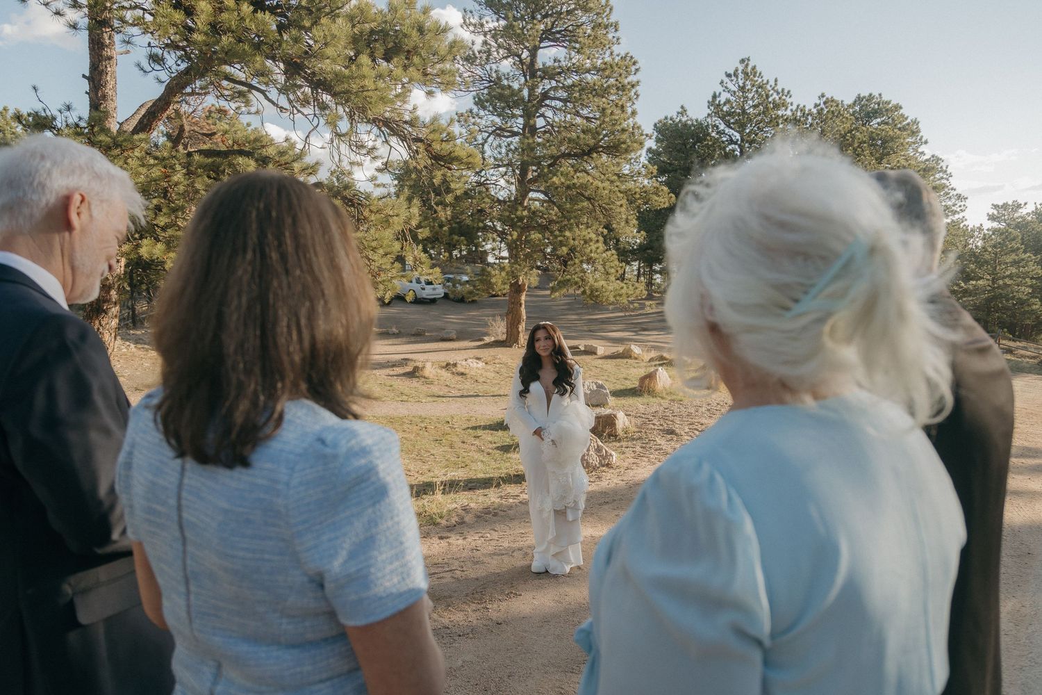 Wedding party listens as officiant speaks at an outdoor ceremony surrounded by pine trees.