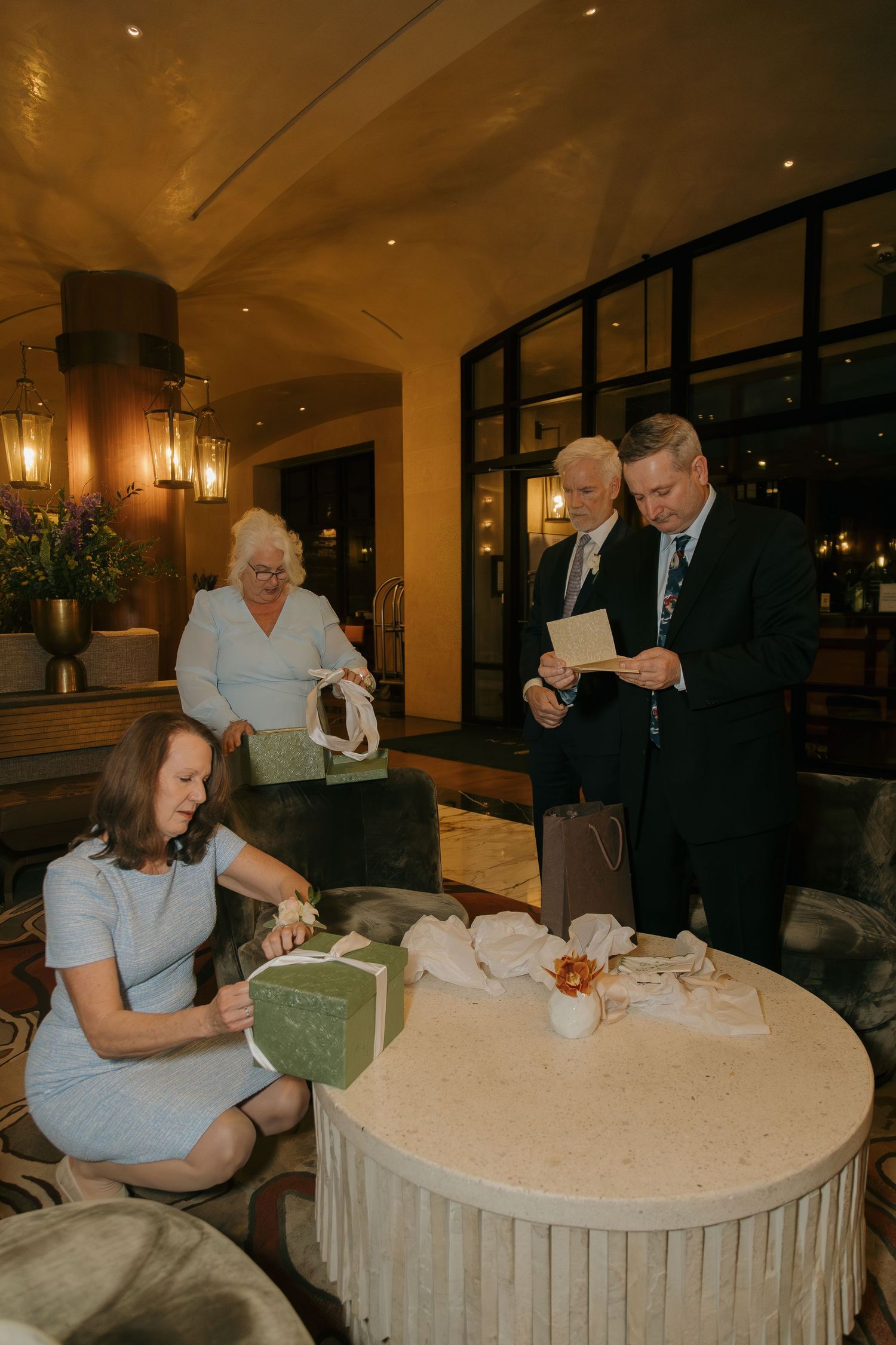 Elegant wedding guests opening gifts at a round table in a warmly lit hotel lobby.