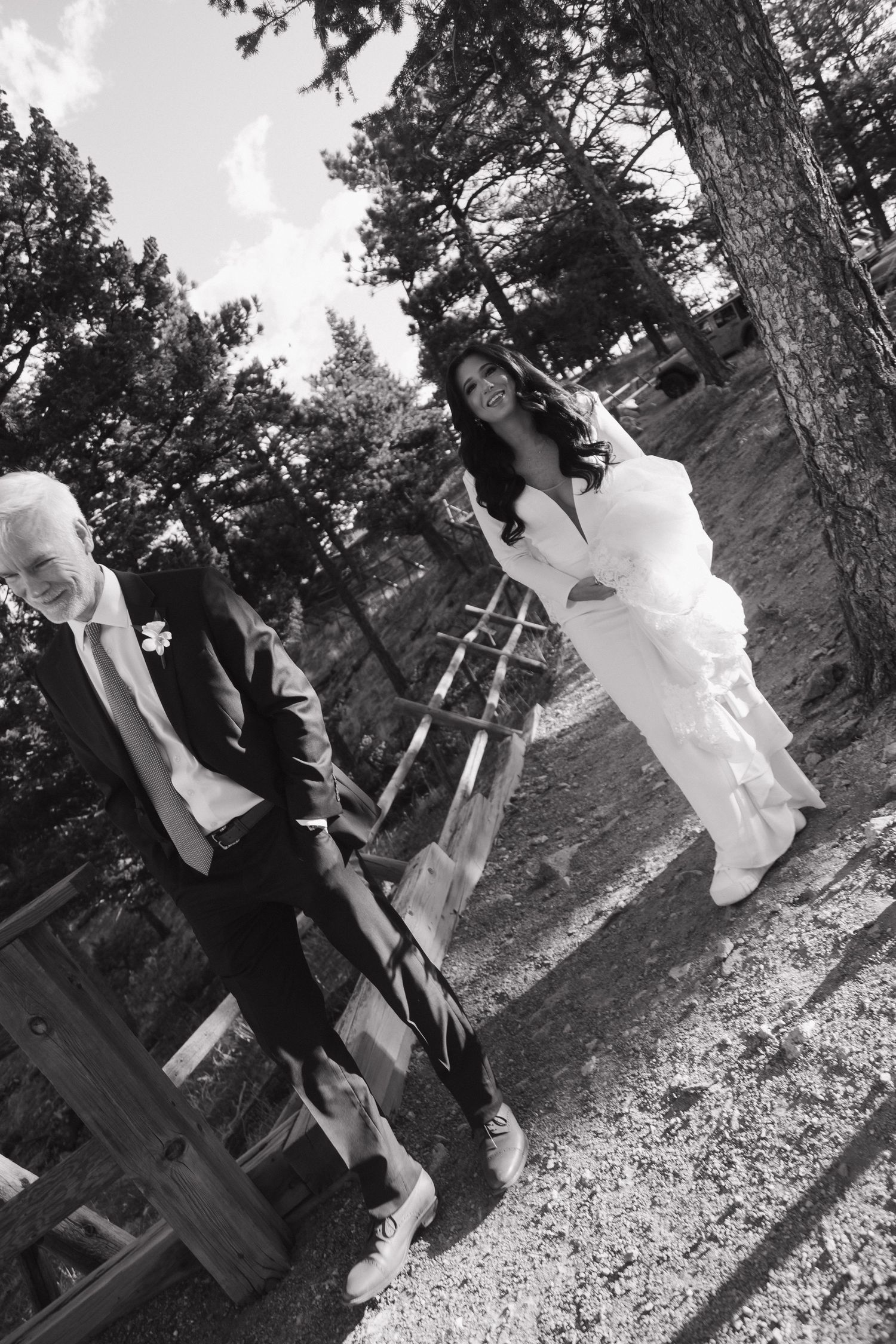 Black and white photo of bride and groom playing on a wooden swing set among pine trees at their outdoor wedding.