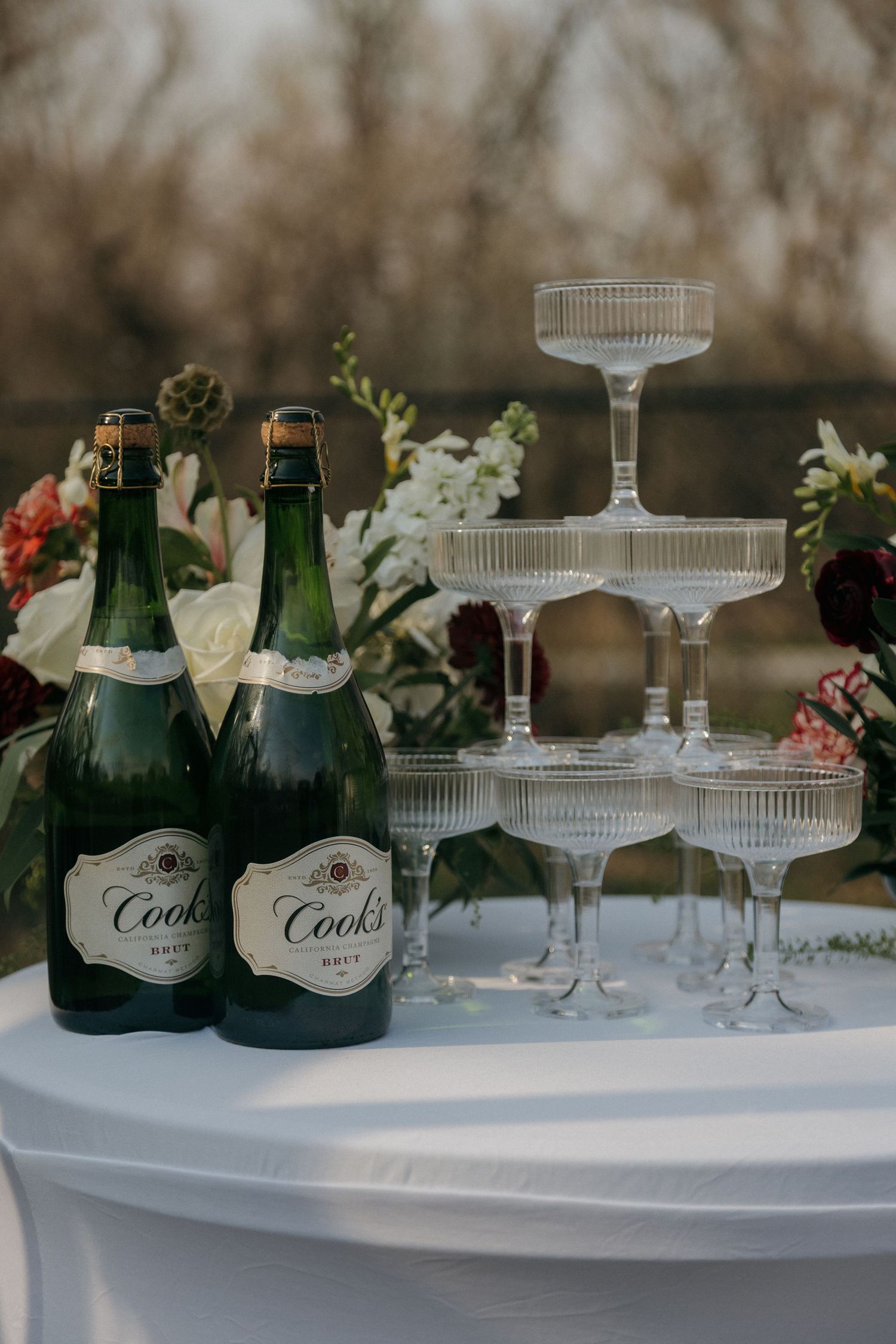 Two Cook's champagne bottles beside crystal coupe glass tower at outdoor wedding reception table.