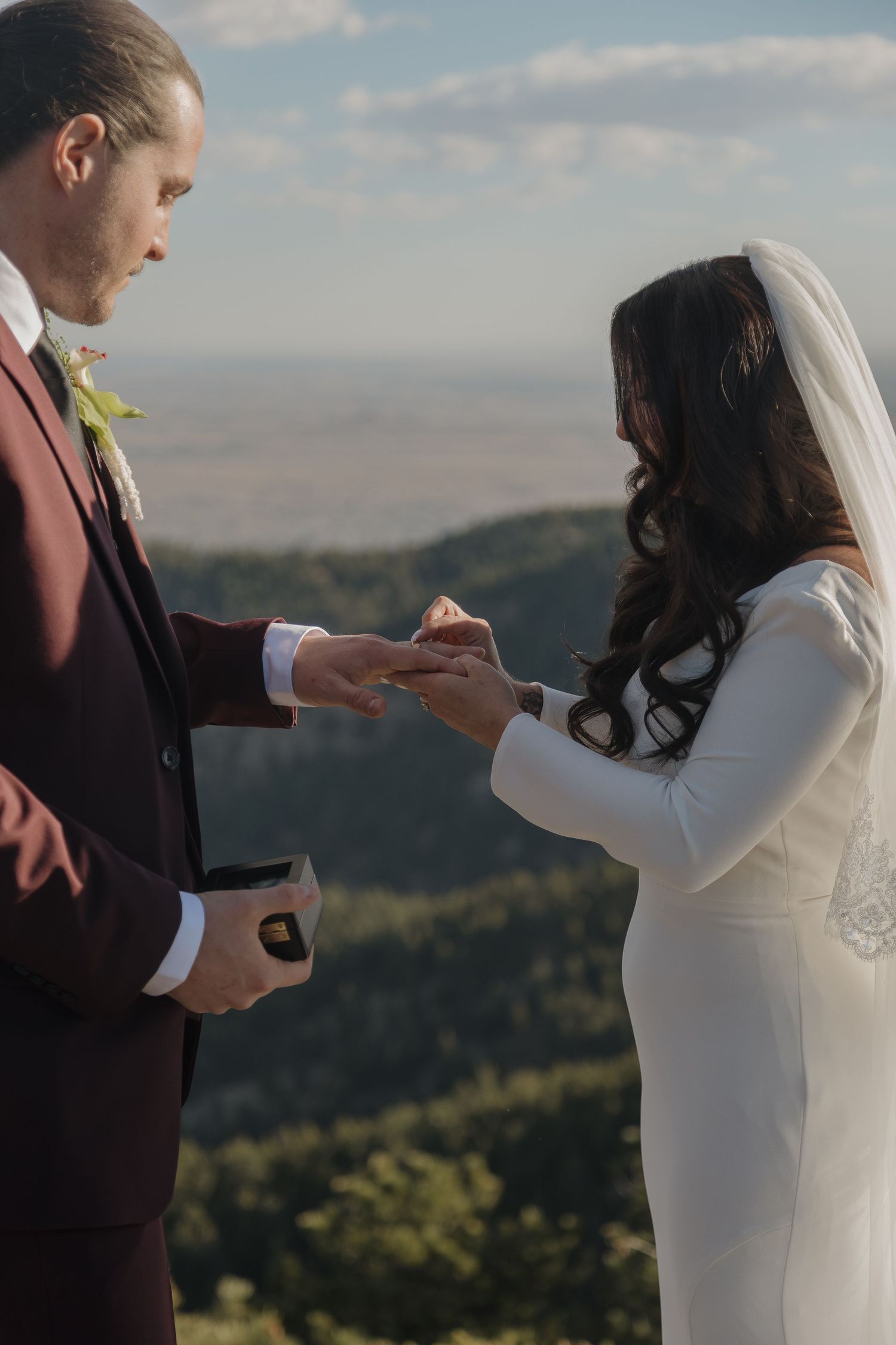 Bride in white gown receives wedding ring from groom in burgundy suit at scenic mountain overlook.