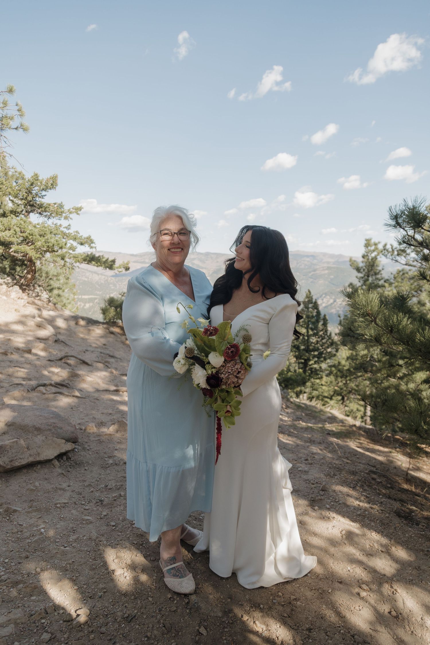 Bride in white long-sleeve gown poses with older woman in blue dress holding bouquet outdoors.