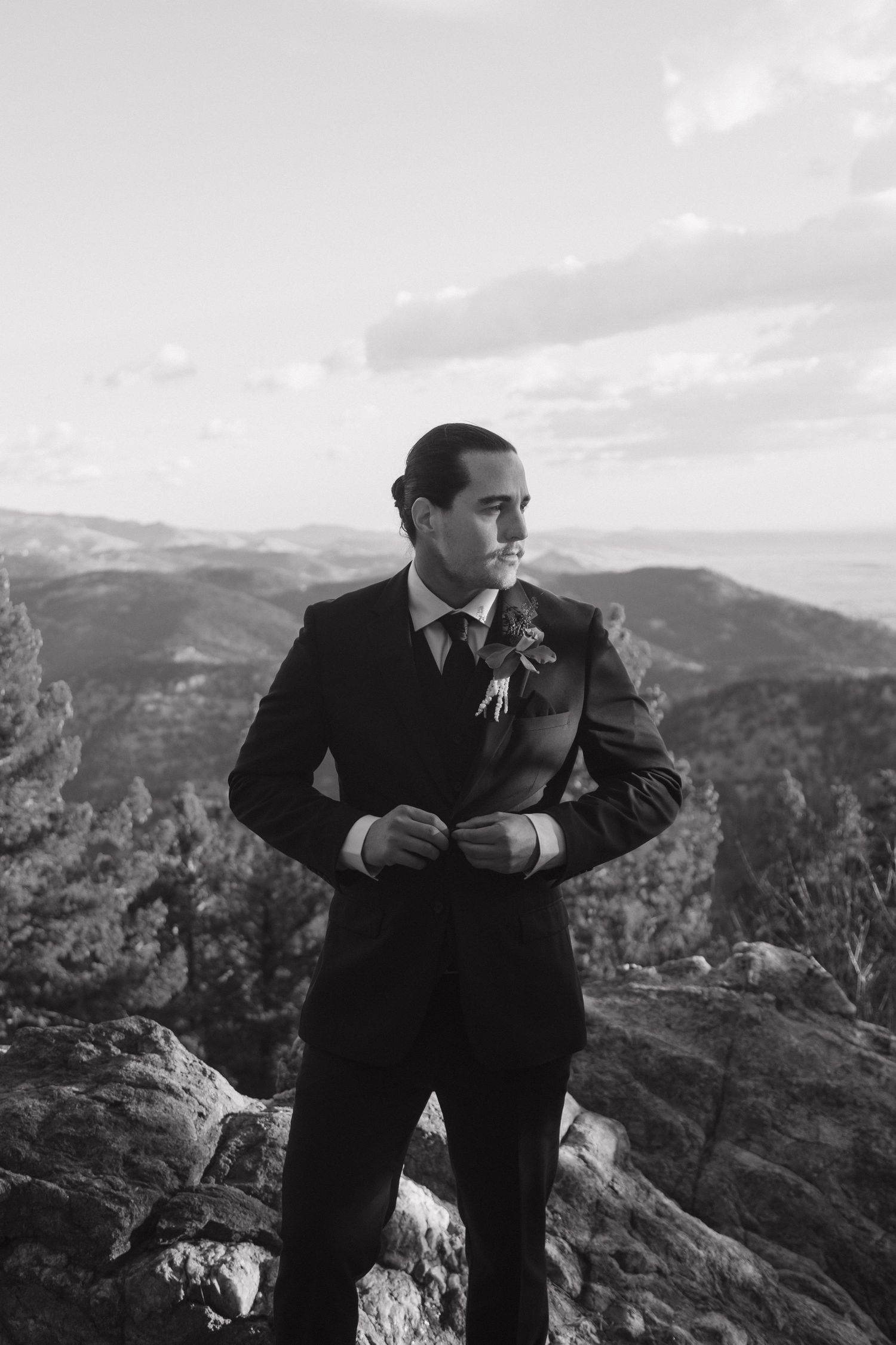 Handsome groom in a dark suit with boutonniere standing on a mountain overlook in black and white photo.