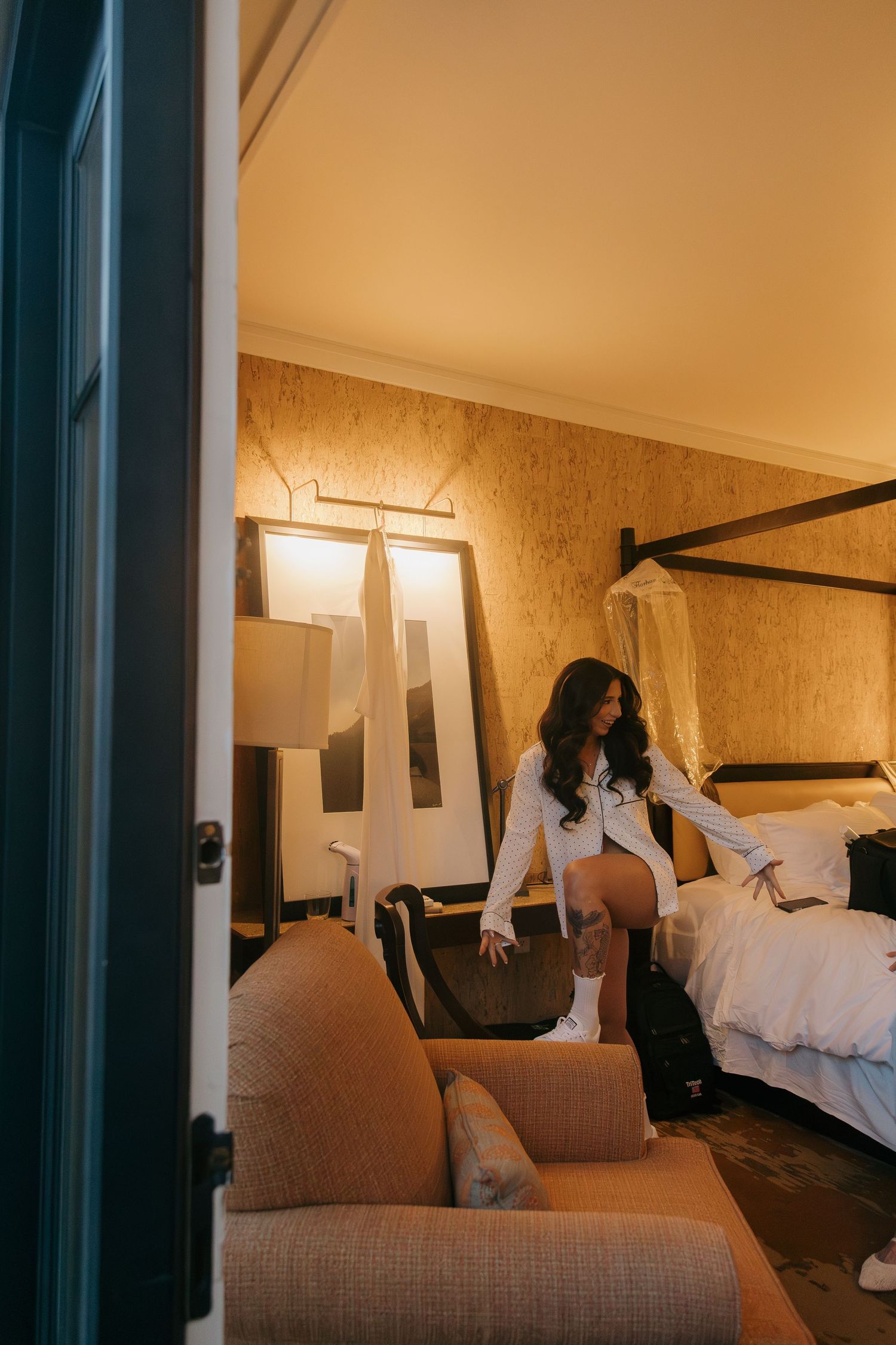 Bride in white robe preparing in warmly lit hotel room with wedding dress hanging nearby.