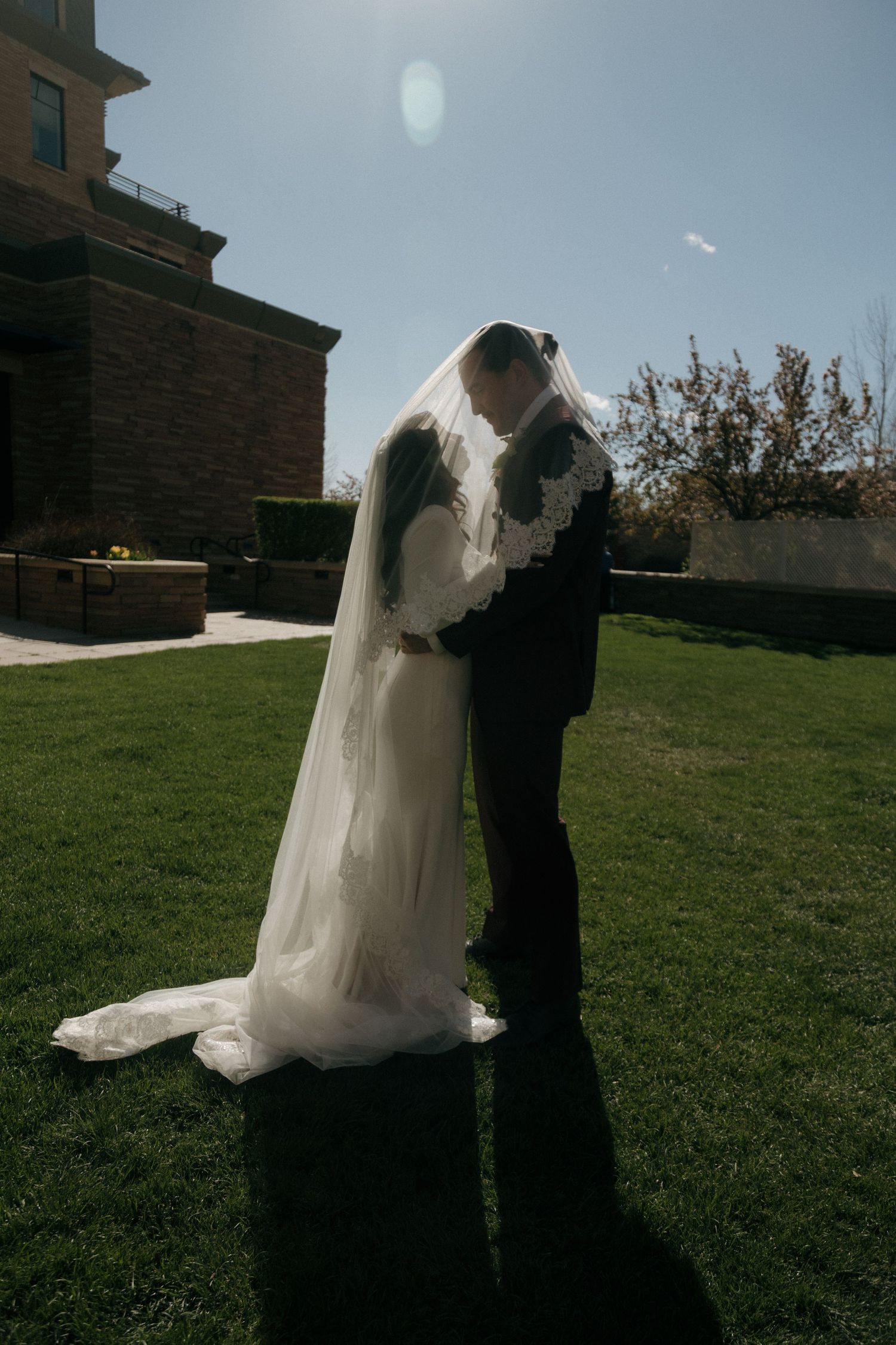 Bride and groom sharing kiss under lace-trimmed veil on sunny green lawn beside stone building.
