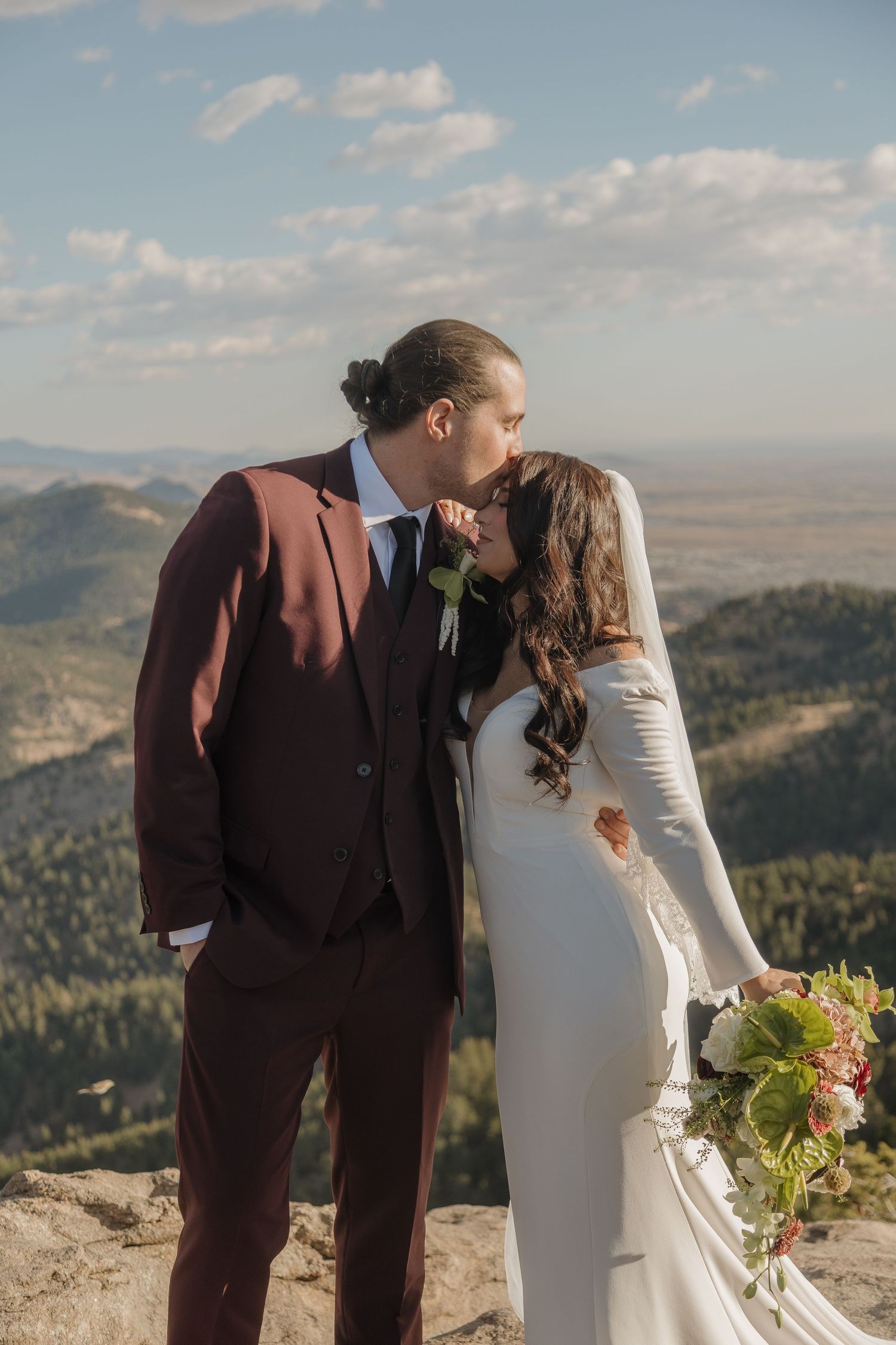Bride and groom share first kiss in burgundy suit on rocky Colorado mountain summit with bouquet in hand.